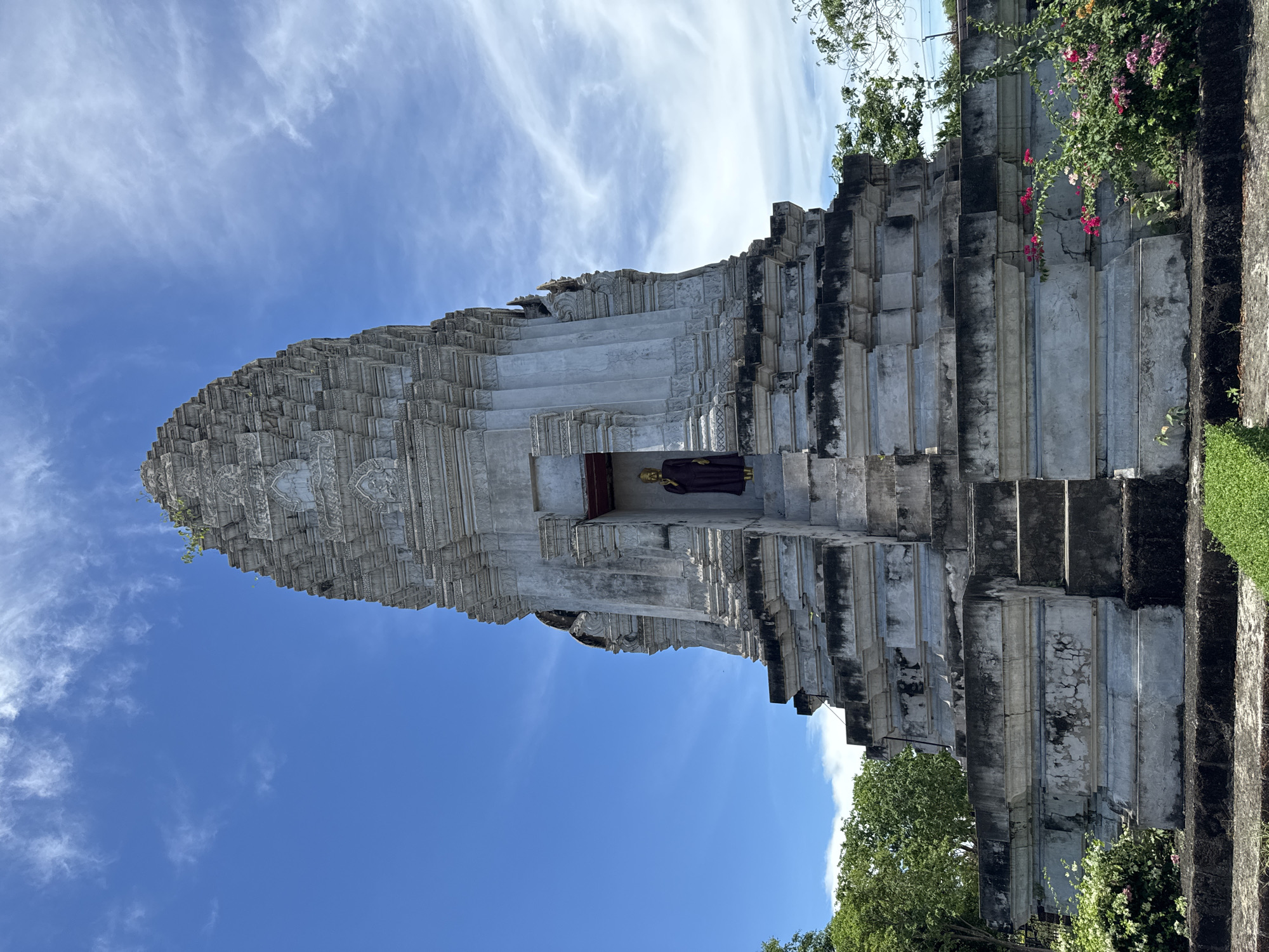 The image shows a tall, white, tiered structure, possibly a temple or pagoda, under a bright blue sky with wispy clouds. The structure is adorned with intricate carvings and has a seated golden statue visible in a central alcove. The architecture includes numerous steps and layered platforms. Vegetation, including flowering plants and green grass, grows around the base of the structure. The location is in the city of Bang Pu Subdistrict Municipality, Thailand.