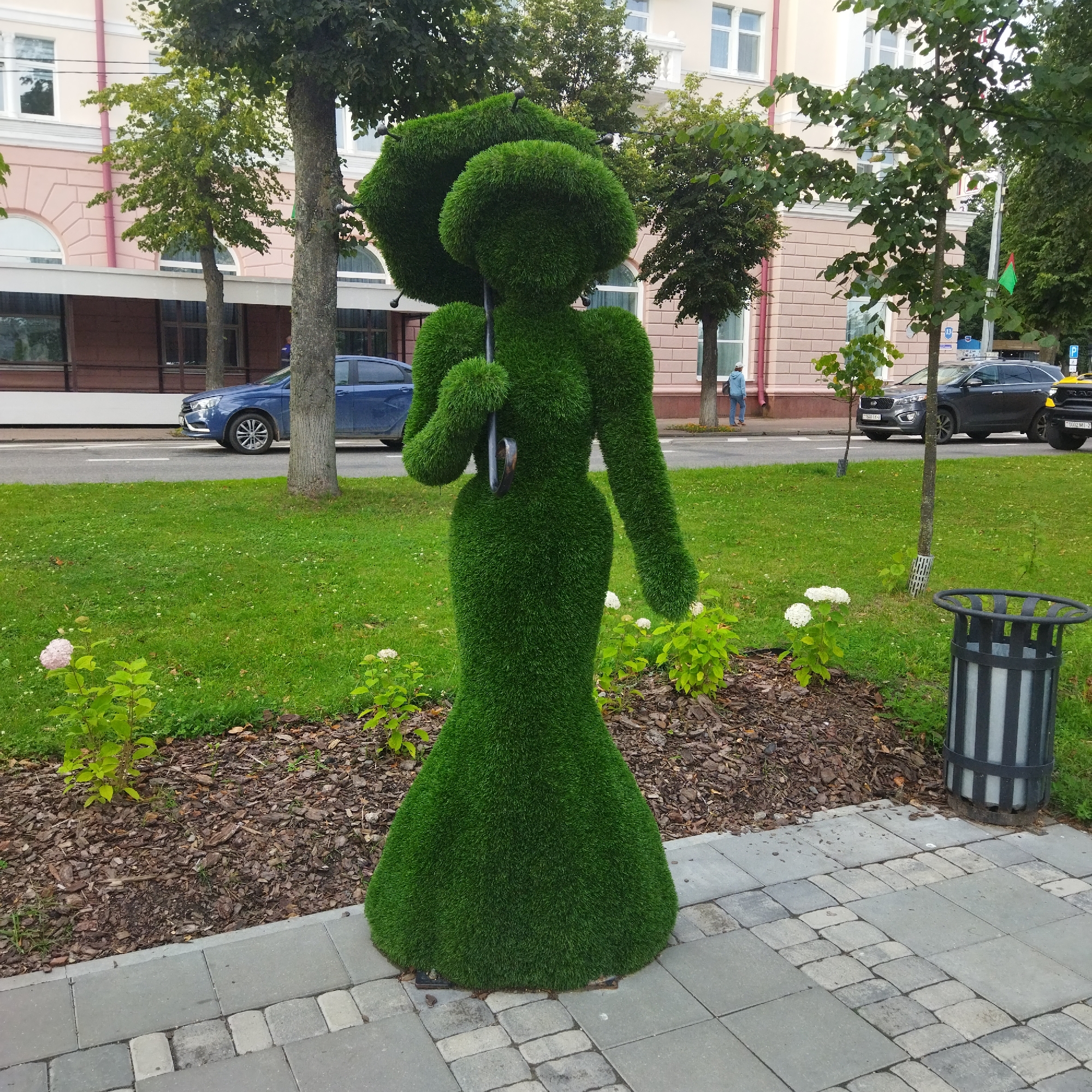 This media file is an outdoor scene at ground level. The primary subject is a topiary in the shape of a woman holding an umbrella. The topiary is made of dense green grass and is positioned on a cobblestone path next to a grassy area with some small white flowers and mulch. Behind the topiary, there's a street with several cars parked and driving. In the background, a building with light pink facade, arched windows, and a white balustrade is visible. Trees with green foliage are scattered throughout the scene. A black trash bin with white vertical stripes stands to the right of the topiary. A person wearing a blue jacket and white cap is walking on the sidewalk in the background. The sky appears overcast, suggesting a cloudy day. The overall impression is a well-maintained urban environment with decorative elements. There are no visible emotions to interpret. The text "1640 189" is visible on the license plate of a dark grey SUV. There is also a green and red flag visible in the distance.