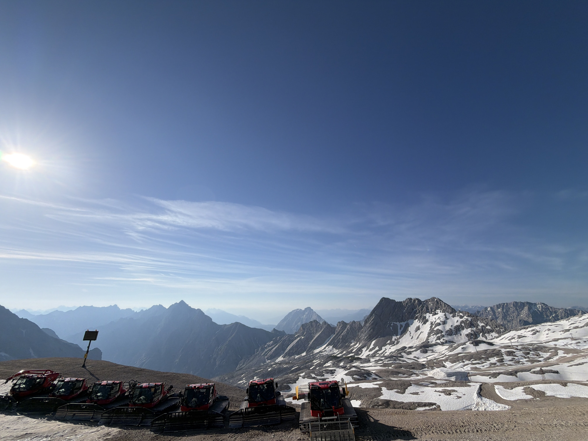 This is a wide, horizontal, eye-level, outdoor photograph taken on a bright, sunny day, likely from a mountaintop. In the foreground, six red snow groomers are parked in a line on a dirt and gravel surface. The snow groomers are pointed towards the left side of the frame. A dark, square sign on a yellow pole is visible behind the first three snow groomers. The middle ground features a vast mountainous landscape with snow-capped peaks and a bright blue sky with wispy clouds. The sun is visible in the upper left corner, casting a bright glare and lens flare. The background reveals layers of distant mountains fading into a hazy blue atmosphere. The overall scene suggests a high-altitude, winter or early spring environment, possibly in the Bavarian Alps near Garmisch-Partenkirchen, Germany, given the presence of snow groomers.