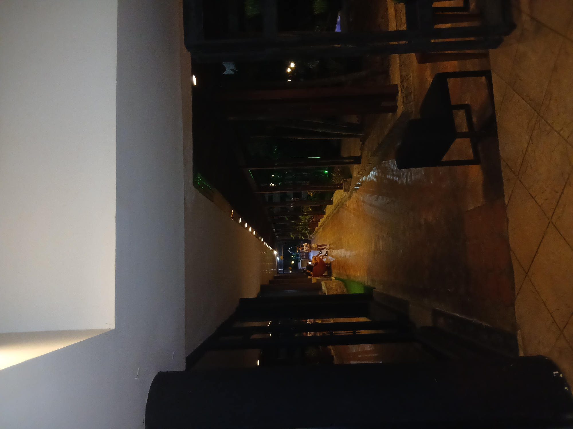 The image captures a long, narrow outdoor space in Playa El Agua, Venezuela. A paved walkway extends into the distance, bordered on one side by a white wall with evenly spaced lights running along its length. On the other side of the walkway is a stone tiled surface. A dark, metal railing and furniture are positioned along this side. People are gathered around tables further down the walkway, suggesting a dining or gathering area. There is a green mat next to a group of people. The overall lighting is dim, with artificial lights illuminating the walkway and highlighting the gathering area. The perspective is slightly angled, adding depth to the scene.