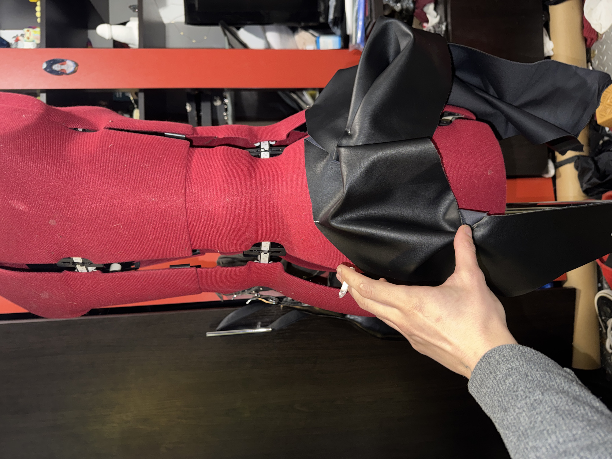 A red fabric upholstered mannequin lies on its side on a dark surface. A hand holding a lit cigarette is near the mannequin. The mannequin appears to be a model for clothing design. Black material drapes over part of the mannequin. The background is cluttered with various objects. The location is a storage or workshop space. The scene is not identifiable as specifically within the city of Sofia, Bulgaria; there are no obvious landmarks or details to confirm this location.