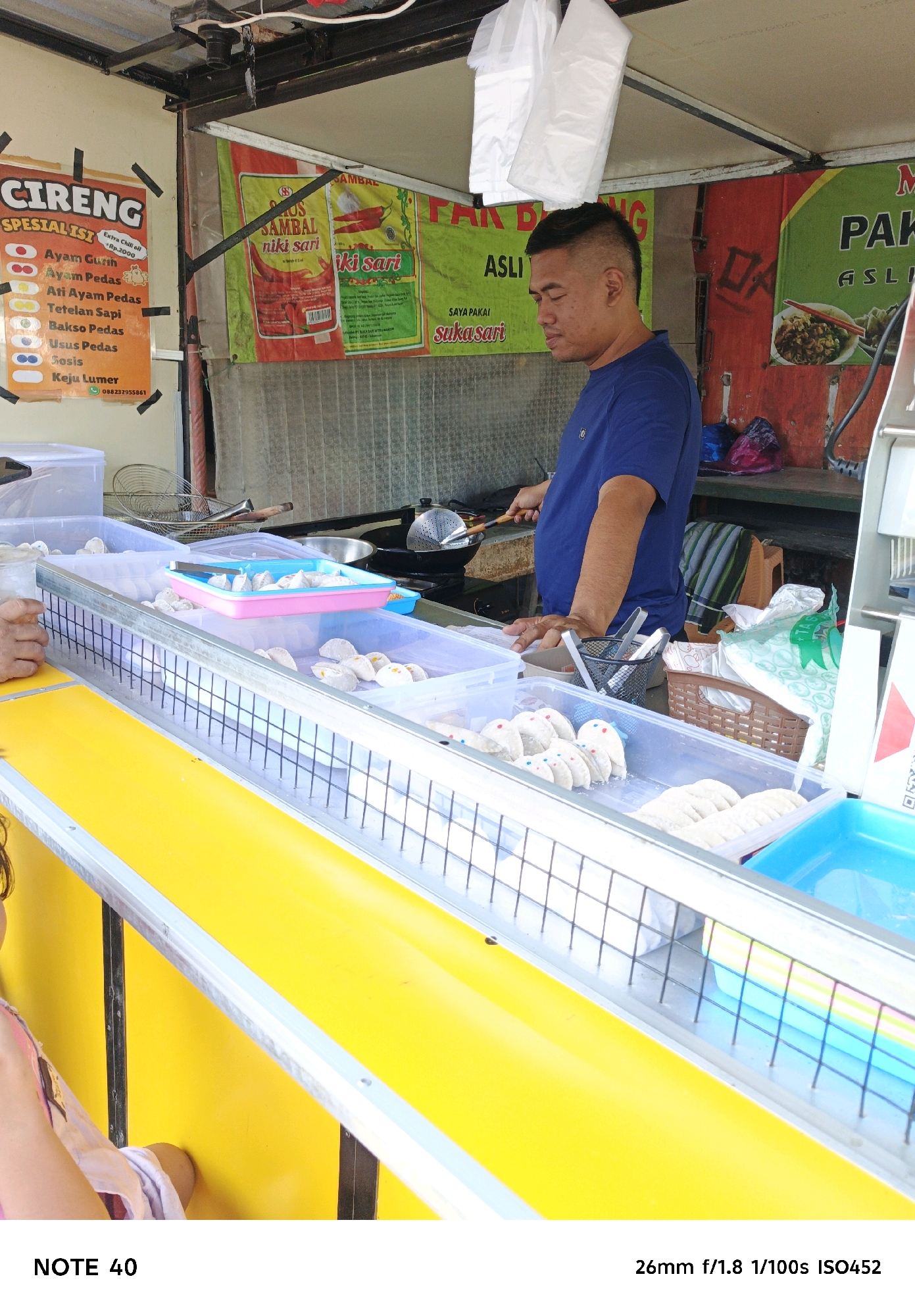 In the city of Kelurahan Cilacap, Kecamatan Cilacap, Indonesia, an outdoor food stall is depicted. The stall features a bright yellow counter and a metal frame displaying various food items in clear plastic containers. The food items appear to be small, white dumplings or fried snacks, some of which are topped with colorful dots. In the background, a man in a blue t-shirt is cooking, with a wok and cooking utensils visible. Banners with Indonesian text and graphics are hung behind the stall, promoting food products. The scene suggests a busy, commercial setting for street food.