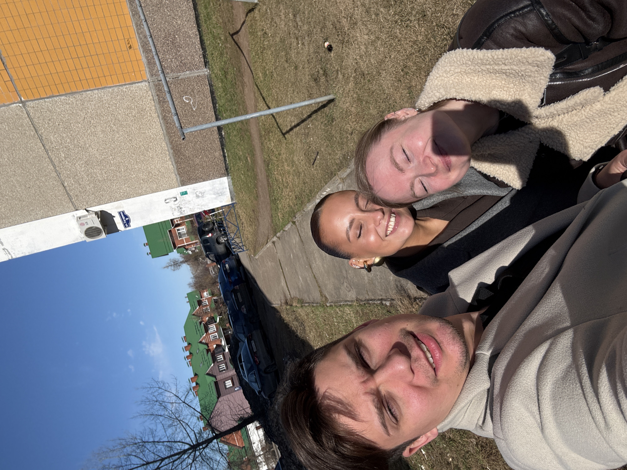 Three people are posing for a selfie outdoors. The man in the foreground wears a white hoodie. The woman in the middle is smiling and wearing a brown top. The woman in the background wears a brown leather jacket with a cream-colored fur lining. Behind them, there is a grassy area, a sidewalk, parked cars, and houses with green roofs. The sky is blue. The photograph was taken in the city of Chaikovsky, Russia, as indicated by the street sign "ул. Декабристов, 5".