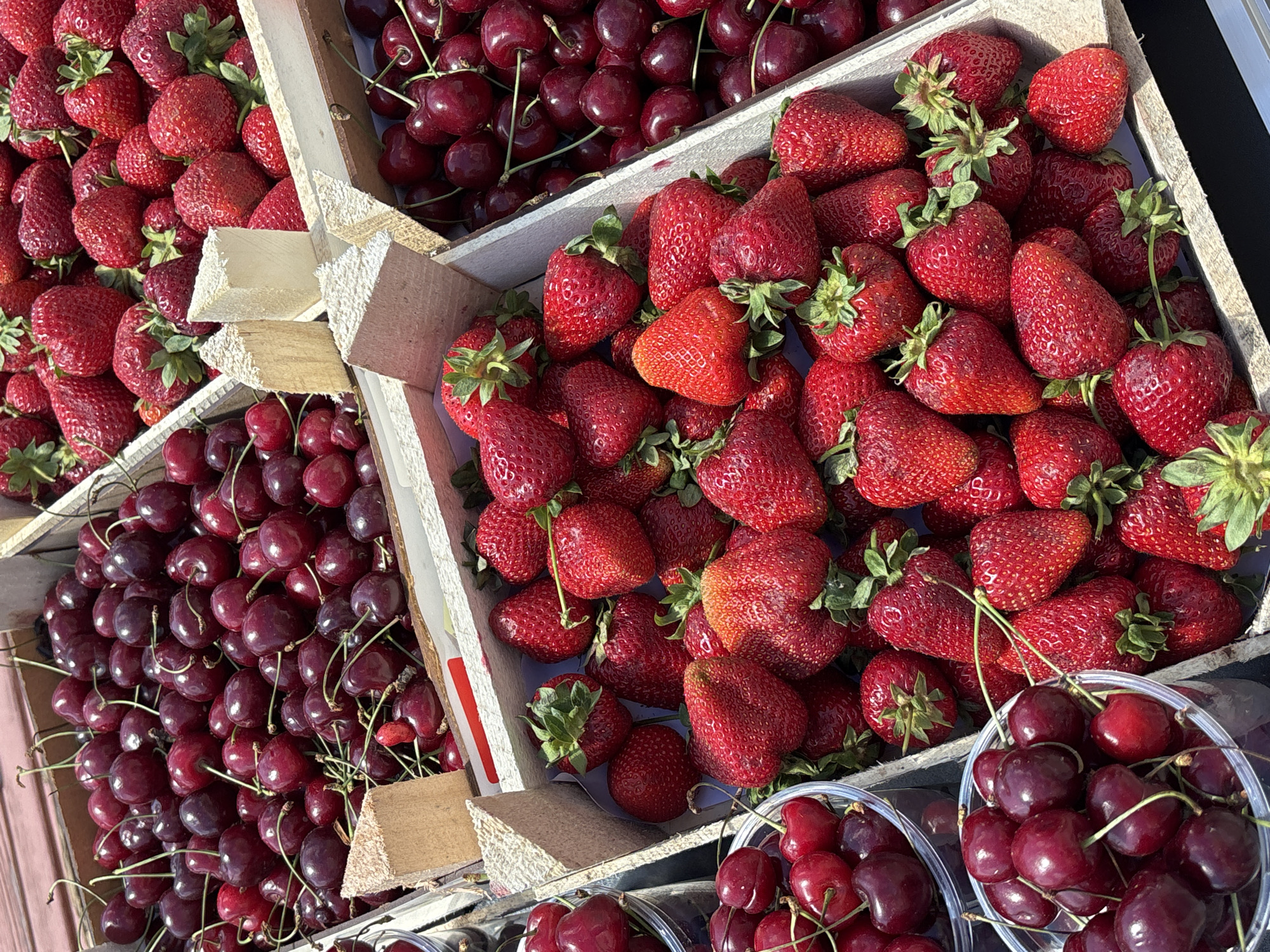 The image shows an outdoor market scene in daylight, possibly in the city of Krasnoyarsk, Russia. There are wooden crates and plastic containers filled with fresh produce, specifically strawberries and cherries. The strawberries are red, ripe, and have green stems attached. The cherries are dark red and also have stems. The crates appear to be handmade and rustic, while the cherries are also in clear plastic containers. The produce is densely packed in the containers, suggesting abundance and freshness. The focus of the image is on the textures and colors of the fruits and the packaging. There are no people visible in the frame.