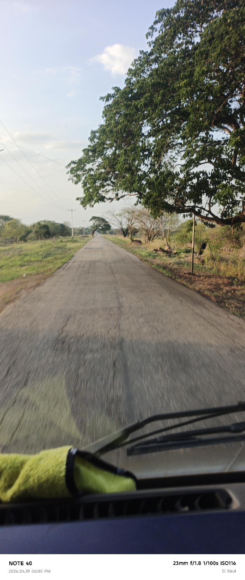 The photograph, taken from inside a vehicle, shows a road stretching into the distance. The road is gray and cracked, with blurred markings indicating motion. Trees line both sides of the road, creating a canopy effect in some areas. A utility pole stands on the left side of the road. The sky is visible between the trees, with soft, pale blue hues. In the foreground, the dashboard of the vehicle is visible, including the windshield wipers and a green cloth. The photo's timestamp indicates it was taken on April 19, 2026, at 6:00 PM in El Baul, La Galera, Venezuela. The camera settings are 23mm f/1.8, 1/100s ISO116.