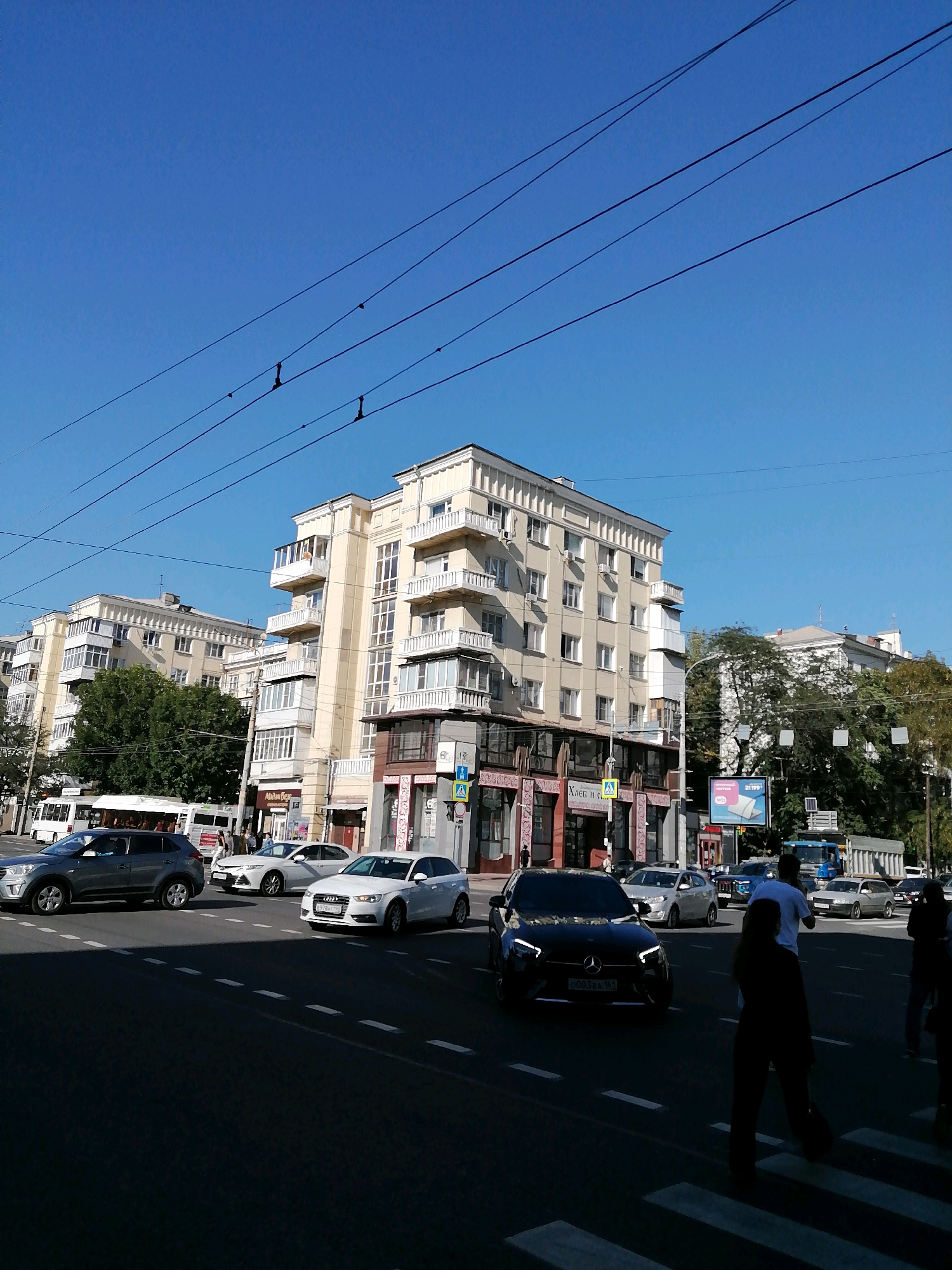 The image depicts a street scene in Rostov-on-Don, Russia. In the foreground, a dark sedan is driving on the road, and a pedestrian is crossing a street. The background features a large, light-colored apartment building with multiple balconies and windows. Several other cars and a bus are visible on the road, and more buildings and trees line the street. Overhead, power lines stretch across the clear blue sky.