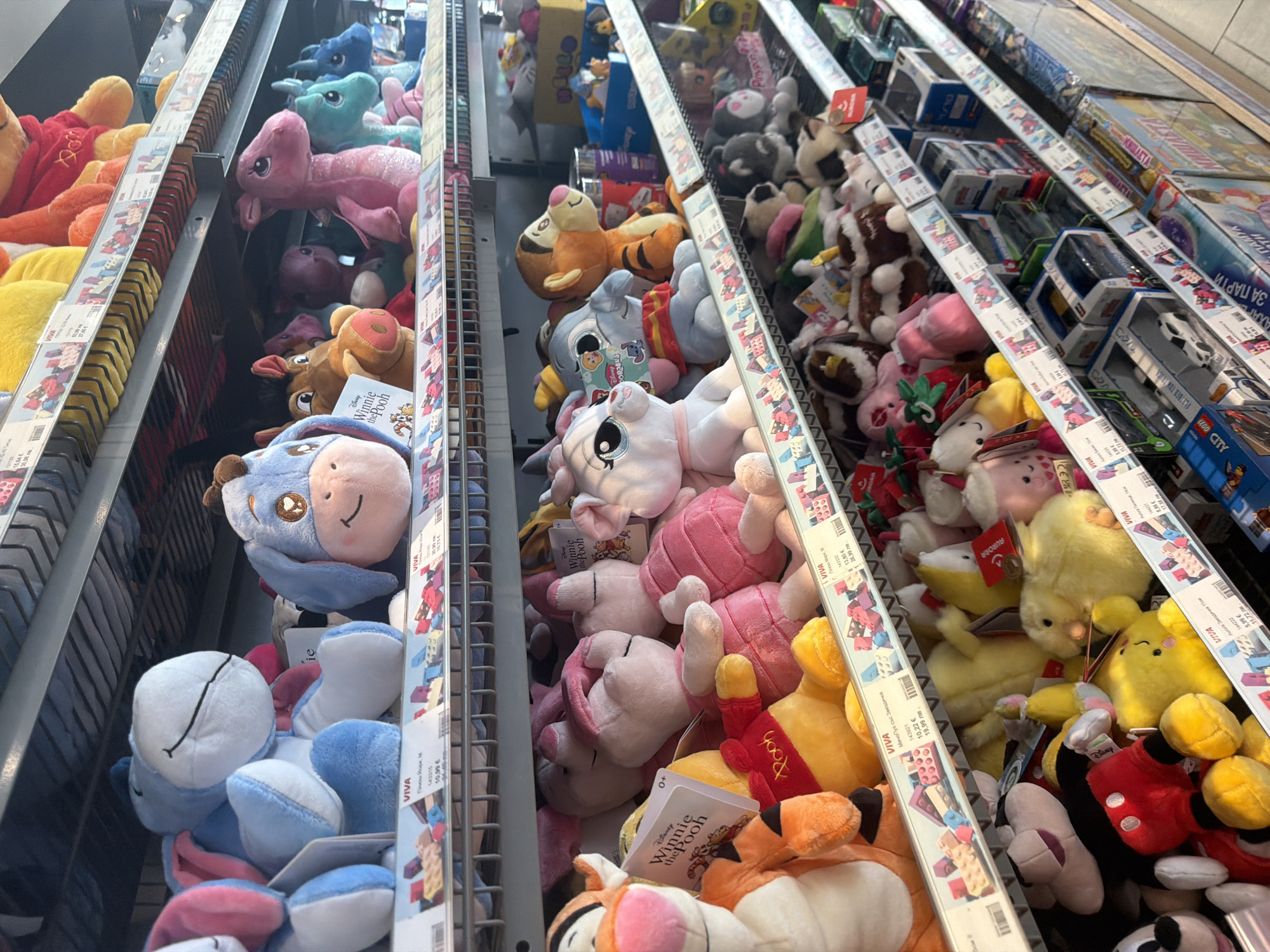 The image displays a vibrant and densely packed collection of plush toys arranged on metal shelves in a retail store aisle. The scene is set in Borovo, Bulgaria, as indicated by the Bulgarian text and dual pricing in Euros and Bulgarian Levs.
Numerous plush toys are the main subjects, featuring a wide variety of characters and animals. Prominently visible are Disney characters such as Winnie the Pooh, Tigger, Eeyore, and Piglet, with multiple copies of each. Other recognizable characters include Mickey Mouse and Pikachu. There are also various generic animal plushies, including pink unicorns, blue and green horses, yellow ducks/chicks, and other cuddly creatures in an array of colors like blue, pink, yellow, brown, and white.
The activity depicted is simply the organized display of merchandise within a retail environment, likely a supermarket or a toy store. The shelves are brimming with items, creating a sense of abundance for potential buyers.
Notable details include the bright, artificial indoor lighting. Many toys have price tags attached, which frequently bear the name "VIVA" and display prices in both Euros (e.g., "10,22 €") and Bulgarian Levs (e.g., "19,99 лв."). Visible Bulgarian text on the tags includes product names such as "Мечо Пух със Звездичка" (Winnie the Pooh with Star), "Ашока Звездичка Пом" (Ashoka Star Pom), "Плюш Йорк," (Plush York,), "Плюш Мари, М" (Plush Marie, M), and "Aura Пиленце 14" (Aura Chick 14). Brand names like "Disney Winnie the Pooh" and "AURORA" are also present. On the upper shelves, packaged toys and boxes, including what appears to be LEGO products (e.g., "LEGO City"), are visible, with some boxes also featuring text like "ЗА ПАРТИ" (For Party). Some tags show an age rating of "+0".