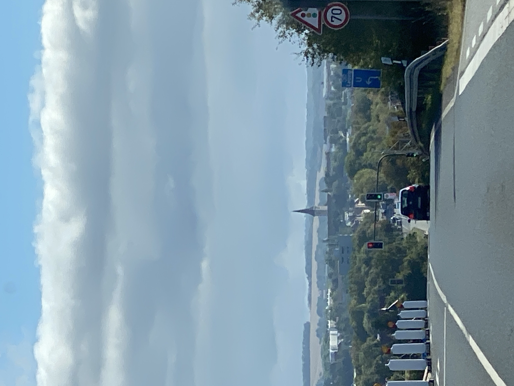 The image shows a view of the city of Werther, Germany, from a road. In the foreground, there is a two-lane road with white markings. On the left side of the road, there are several white temporary traffic barriers. On the right side of the road, there are traffic signs, including a speed limit sign indicating 70 and a traffic light ahead.
In the middle ground, there is a dark-colored car driving on the road. Further in the background, the city of Werther is visible, with trees, buildings, and a prominent church steeple. The sky is partly cloudy. The overall scene depicts a typical view of a city from a road leading into it.