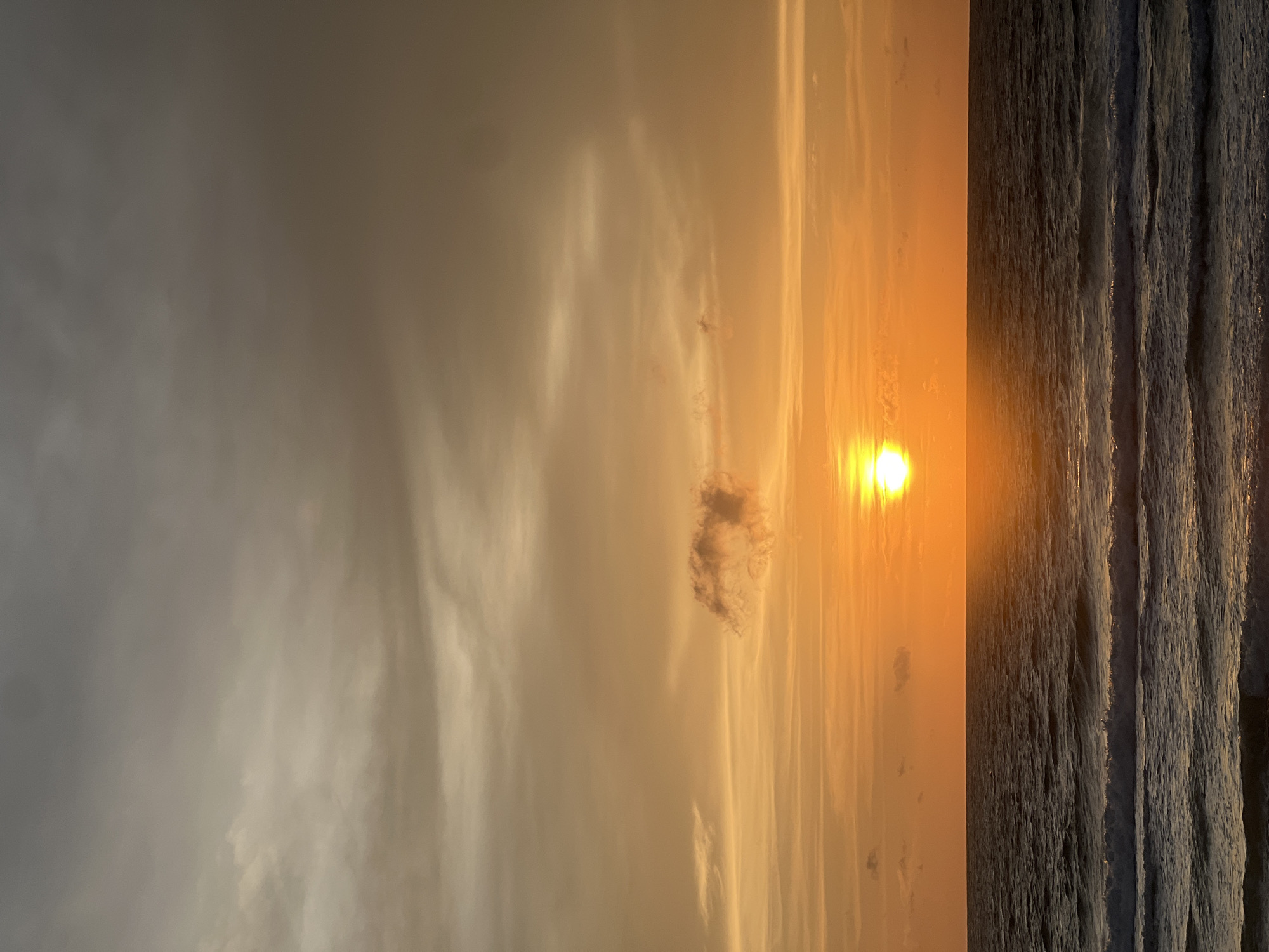 The image depicts a serene ocean sunset in Choeng Thale, Thailand. The sun, a bright, glowing orb, is positioned just above the horizon, casting a warm, golden hue across the sky. The sky is filled with a gradient of colors, ranging from deep orange near the horizon to a softer, muted grey at the top. Wispy clouds are scattered throughout the sky, some catching the sunlight and appearing as streaks of pale gold.
The ocean in the foreground is calm, with gentle waves rolling towards the shore. The water reflects the colors of the sunset, creating a shimmering, iridescent surface. The horizon line is sharp and distinct, separating the vast expanse of the sea from the colorful sky. There are no people or man-made objects visible in the scene, emphasizing the natural beauty of the environment. The overall impression is one of tranquility and natural splendor, characteristic of a coastal sunset in Thailand.