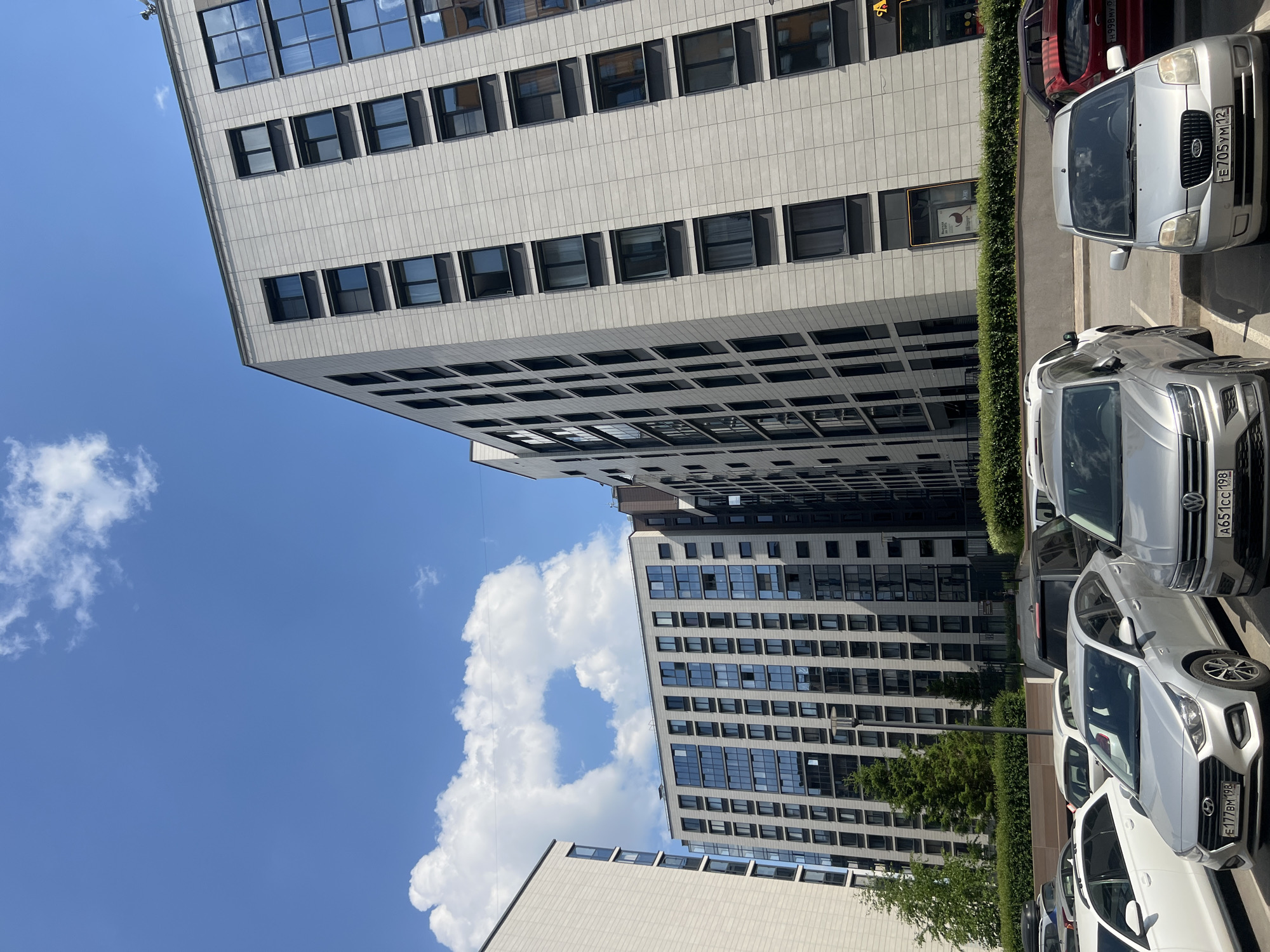 The image, correctly oriented, presents a vibrant, sunlit urban scene in Saint Petersburg, Russia, characterized by modern architecture and parked vehicles.
In the foreground, a line of cars is parked parallel to a lush green hedge. The closest car is a silver Kia Picanto, bearing the Russian license plate E05YM12. Behind it, a silver crossover or SUV is visible with the license plate A651CC198. Further along the row, another silver car displays the plate E177BM198. A red car is partially visible behind the Kia. The hedge creates a neat boundary between the parking area and the buildings.
The midground features two prominent, tall buildings. The closest building is a light-colored, possibly off-white or light gray, high-rise structure. Its facade is composed of a regular grid of large, rectangular windows, many of which reflect the bright blue sky and clouds. Another similar modern building can be seen slightly further back and to the right, also featuring numerous windows. The architectural style is contemporary and functional.
The background is dominated by a clear, bright blue sky. Scattered across the sky are several distinct, fluffy white cumulus clouds, suggesting a sunny and pleasant day. The bright daylight casts subtle shadows, enhancing the textures of the buildings and the vehicles. The visible license plates, particularly those ending in '198', confirm the location context of Saint Petersburg.