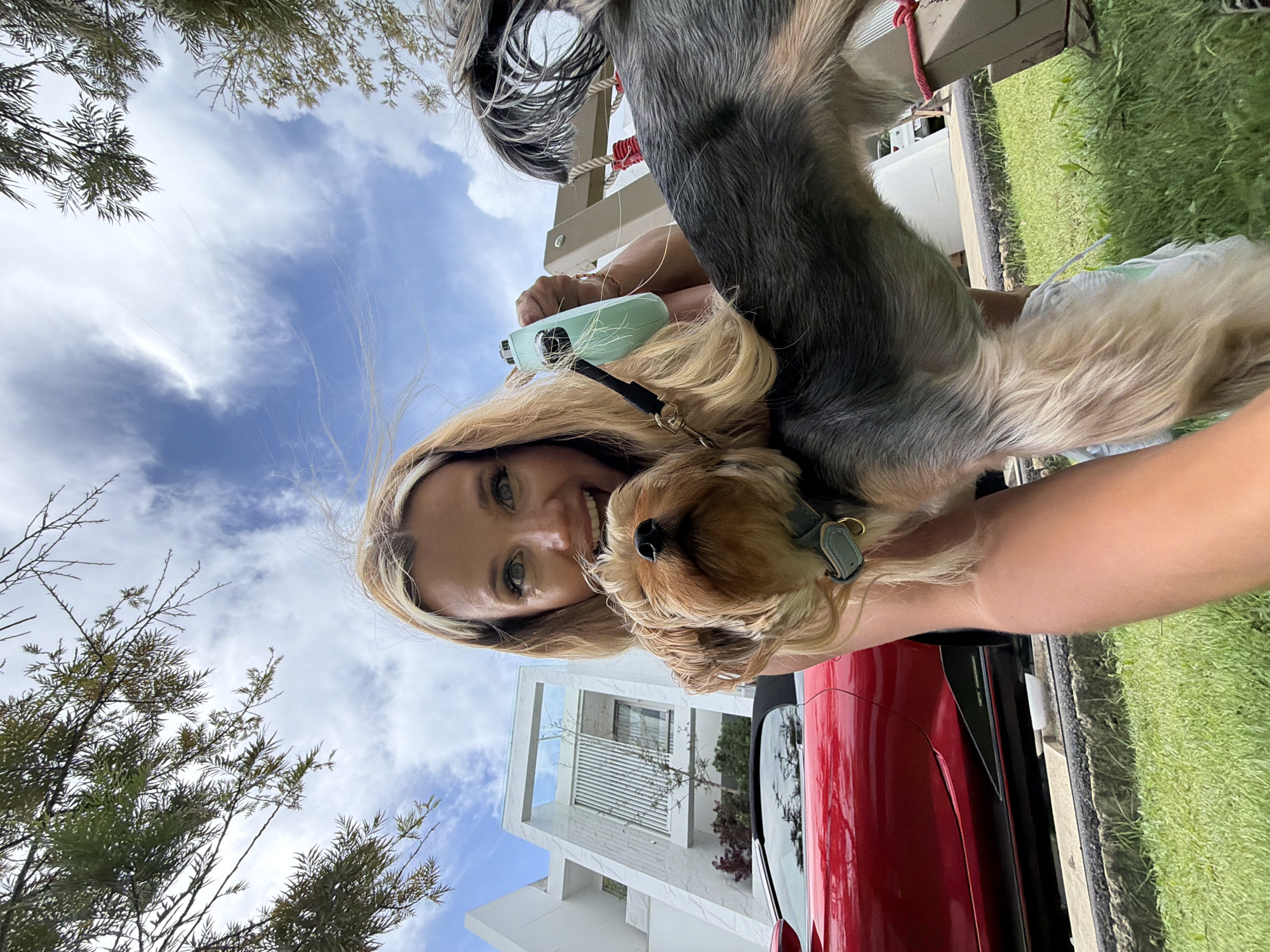 A woman is taking a selfie with her small dog in the city of Thep Krasatti, Thailand. The woman has blonde hair and is smiling at the camera. The dog is a Yorkshire Terrier with brown and grey fur and is looking at the camera. The background shows a modern white house and a red sports car. The scene is outdoors, with grass and trees in the foreground and a blue sky with clouds above. The woman is holding a retractable dog leash in her right hand.