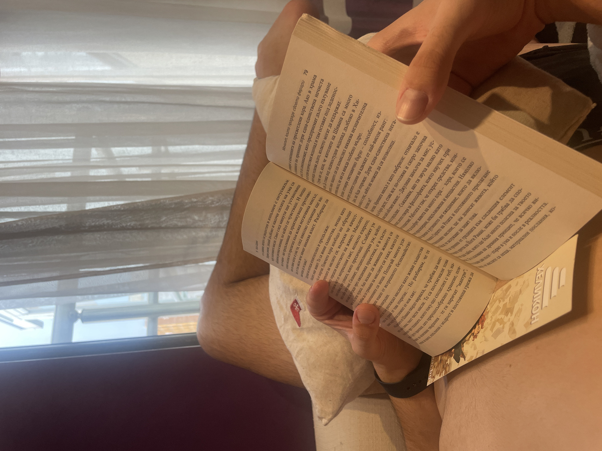 A person sits reading a book in Bulgarian. The book is open to pages 79 and 80, with the title of the book visible as "Азн Шрма" on the spine. The text on the pages includes passages discussing thoughts, enlightenment, and living a fulfilling life. The individual's left hand holds the book open, while their right hand supports it. They are wearing a watch on their left wrist. The person appears to be sitting on a pillow. In the background, a window with white blinds partially obscures the view outside. The image does not show any specific details about the city or location to identify it specifically as Sofia, Bulgaria.