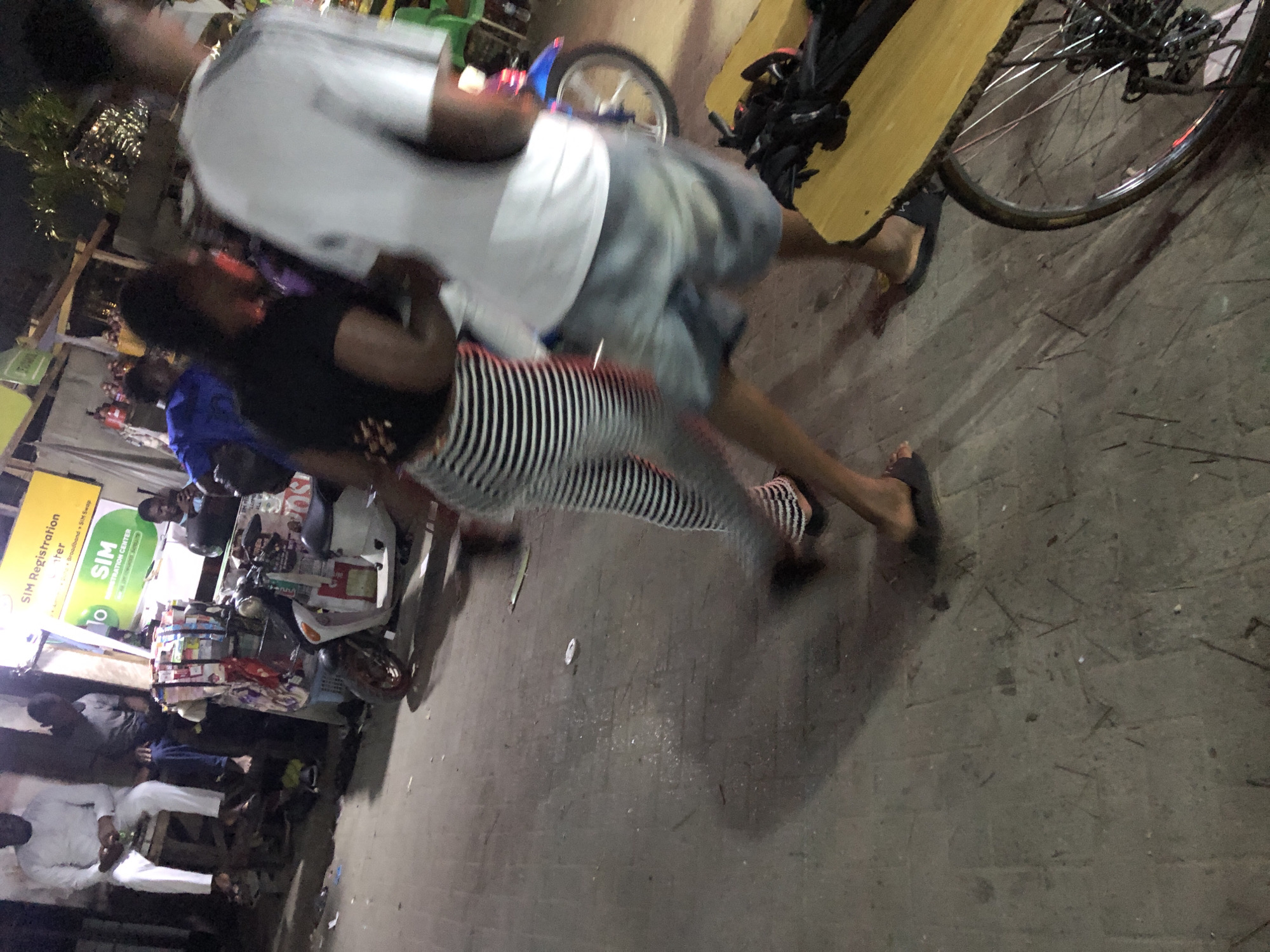 The image, taken at night, shows a street scene in Moba, Nigeria. A woman wearing a black top and black and white striped pants walks alongside a man in a white shirt and blue shorts. They are in front of a "SIM Registration Center" stand that has a sign in green and yellow. In the background are people sitting on chairs, and a motorcycle is visible near the stand. A bicycle is parked to the right of the walking figures. The ground appears to be paved.