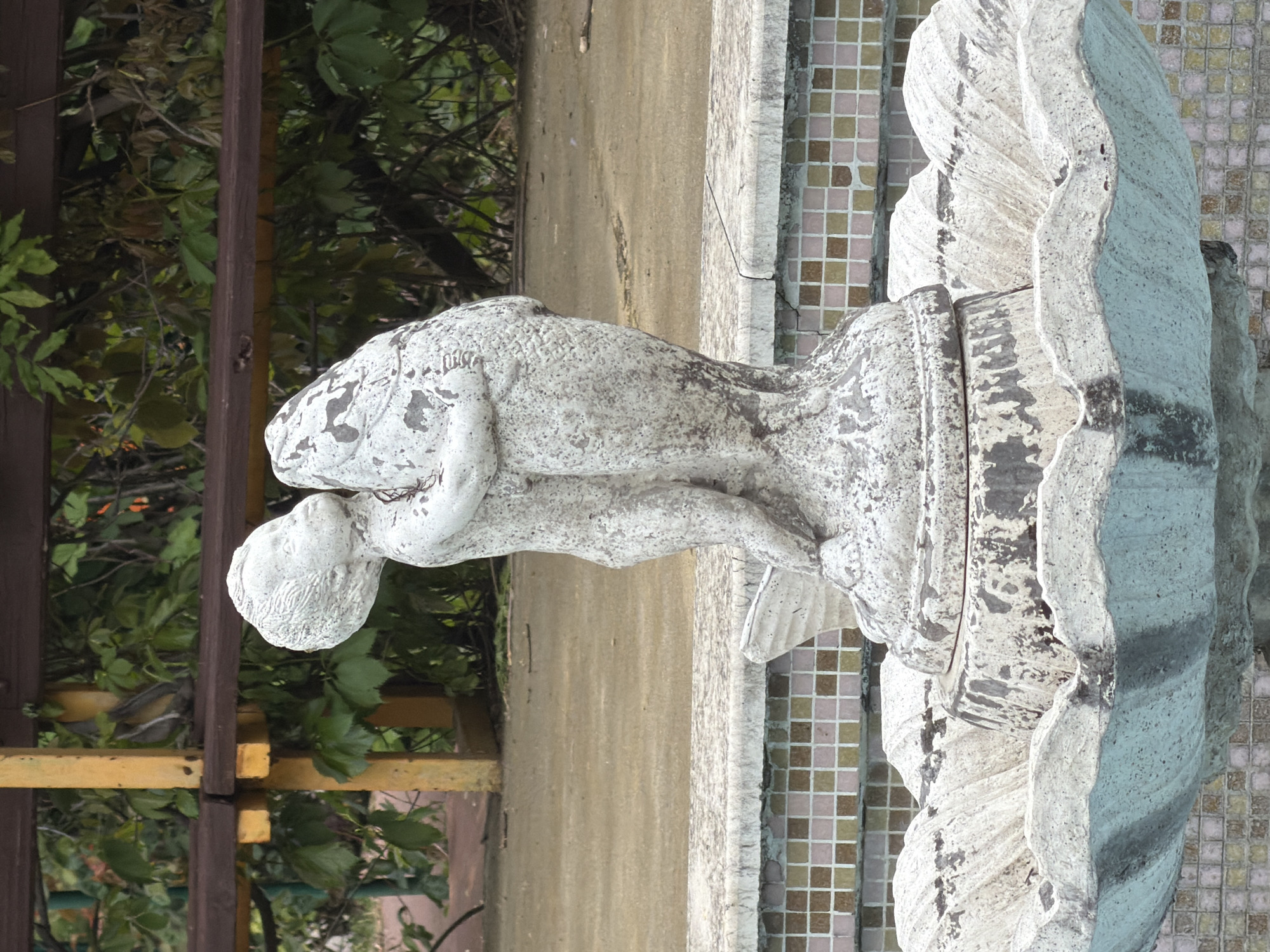 The image features a weathered white stone statue, oriented upright, depicting a putto or cherub-like figure holding a large fish. The statue is a central element, likely part of a fountain or garden ornament, showing considerable signs of age with dark discoloration and chipped surfaces revealing a darker material underneath. The figure has a rounded form, embracing the fish, which has an open mouth and visible scales.
Behind the statue, on the right side, a wall or structure is covered with small, square mosaic tiles in hues of light pink, beige, and brown. To the left and in the background, abundant green foliage from bushes or trees forms a dense natural setting. Within this foliage, dark brown vertical wooden posts and yellow horizontal beams are visible, suggesting a fence, trellis, or pergola. A light-colored concrete or stone surface also runs horizontally across the midground, behind the statue. The scene is outdoors, likely a garden or park in Pazardzhik, Bulgaria, under natural daylight. There is no visible text or human activity.