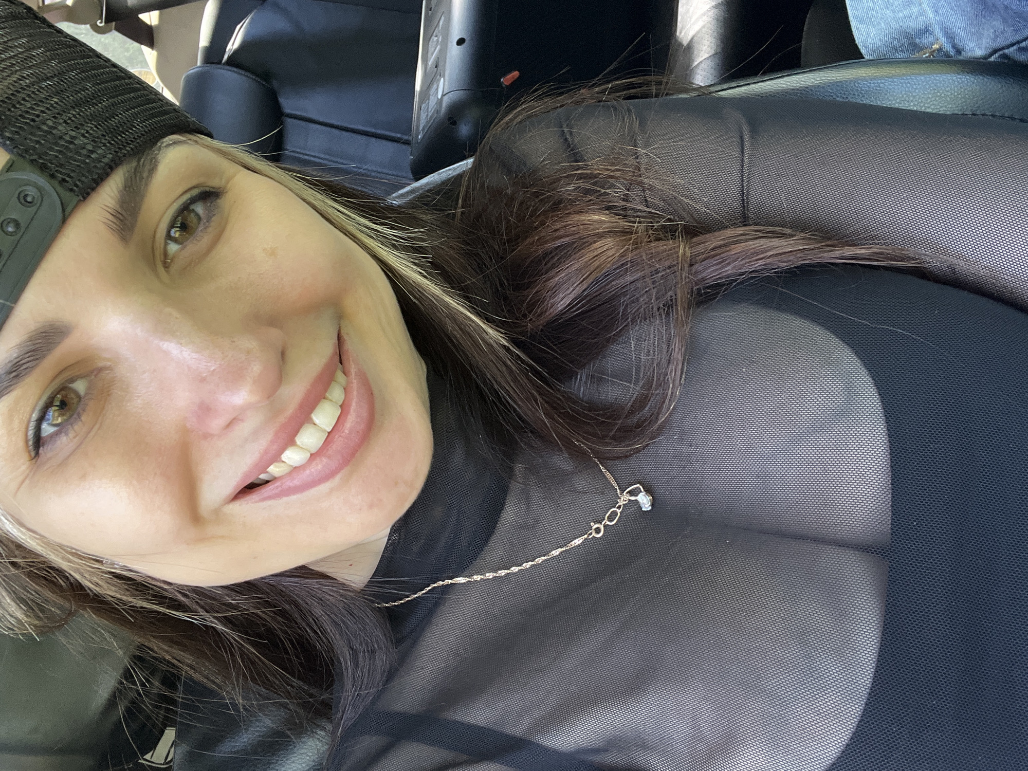 A selfie of a smiling woman with light skin and brown hair with blonde highlights, wearing a black baseball cap and a sheer black top with a silver necklace. The background appears to be the interior of a car, with black leather seats and a glimpse of blue jeans.