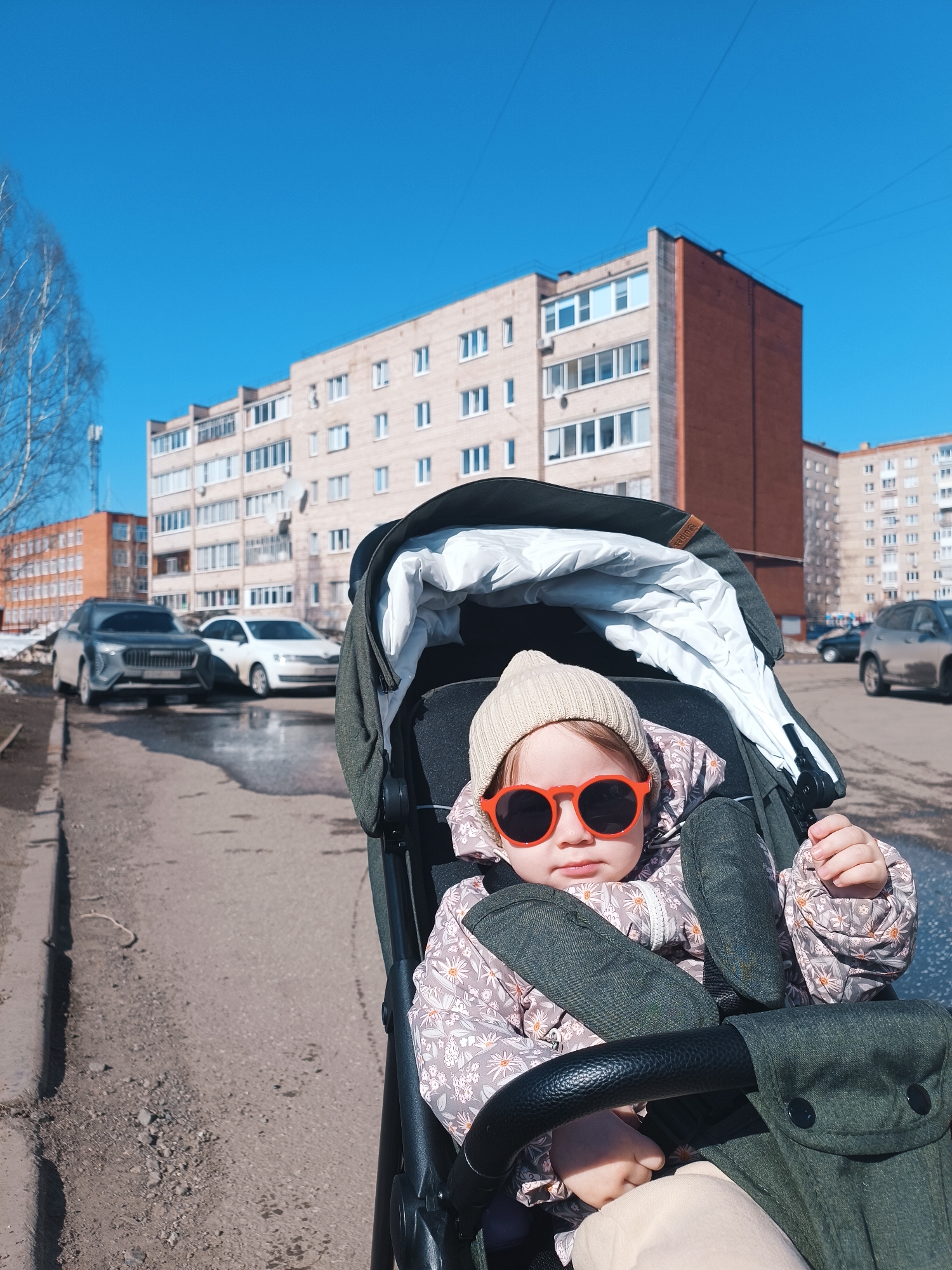A small child wearing a beige hat and round red sunglasses is sitting in a stroller on a paved surface in what appears to be a residential area. The child is wearing a patterned jacket. Behind the stroller, several multi-story brick buildings are visible. There are cars parked on the street, and patches of snow or melting ice are near the curb. The sky is clear and blue. This photo appears to be taken in the city of Izhevsk, Russia.