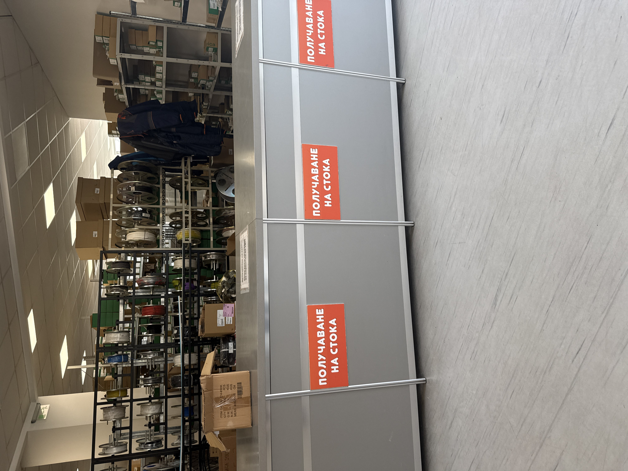 This is a medium-shot, eye-level, interior photograph of a retail or warehouse environment, likely in Sofia, Bulgaria. The scene depicts a service counter with signage indicating "ПОЛУЧАВАНЕ НА СТОКА" (Receiving Goods). The counter is made of gray laminate with vertical silver metal supports.
On the counter, there are several cardboard boxes. One open box on the left displays labels with item details. Behind the counter, rows of shelving units are filled with various items, including spools of wire or cable, and packaged goods. A dark blue jacket is hung on one of the shelves, suggesting a staff area. The floor is a light gray, linoleum-like material with faint streaks. The ceiling features fluorescent lighting panels. The overall impression is that of a functional, commercial space.