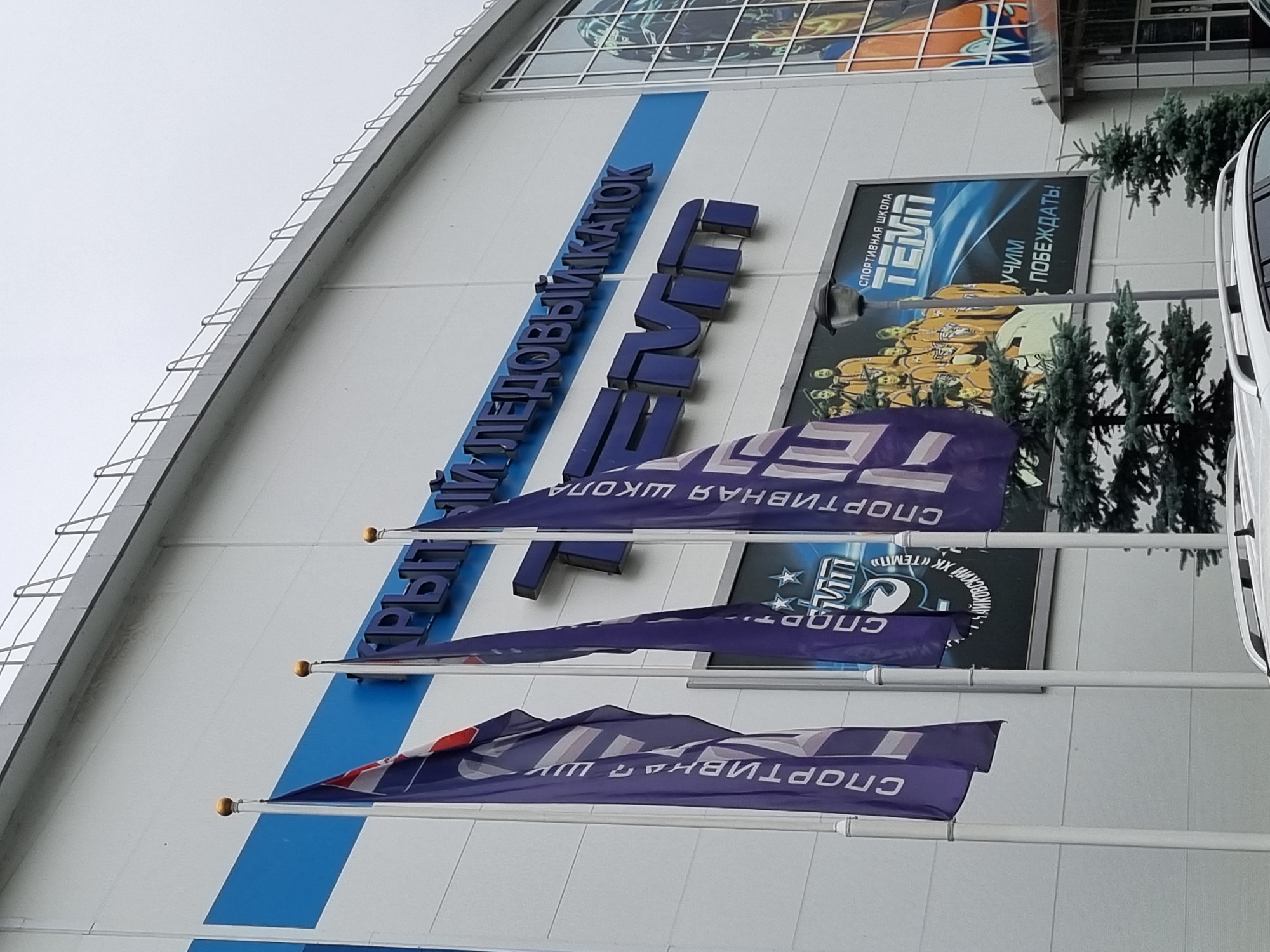 The image shows the exterior of a modern building with the text "КРЫТЫЙ ХОККЕЙНЫЙ КАТОК" and "TEMП" in large blue letters. Three purple flags are displayed in front of the building, each with white text and graphics. One flag appears to have a red and white emblem. To the right of the flags, there is a large advertisement poster featuring a group of people in orange uniforms and the text "СПОРТИВНАЯ ШКОЛА TEMП УЧИМ ПОБЕЖДАТЬ!". The building has a white facade with a blue stripe running horizontally above the text and windows on the right side. Two evergreen trees are visible in front of the building, and a portion of a white car is in the foreground. The sky is overcast. This building is identified as the "TEMП" ice rink in Chaikovsky, Russia.