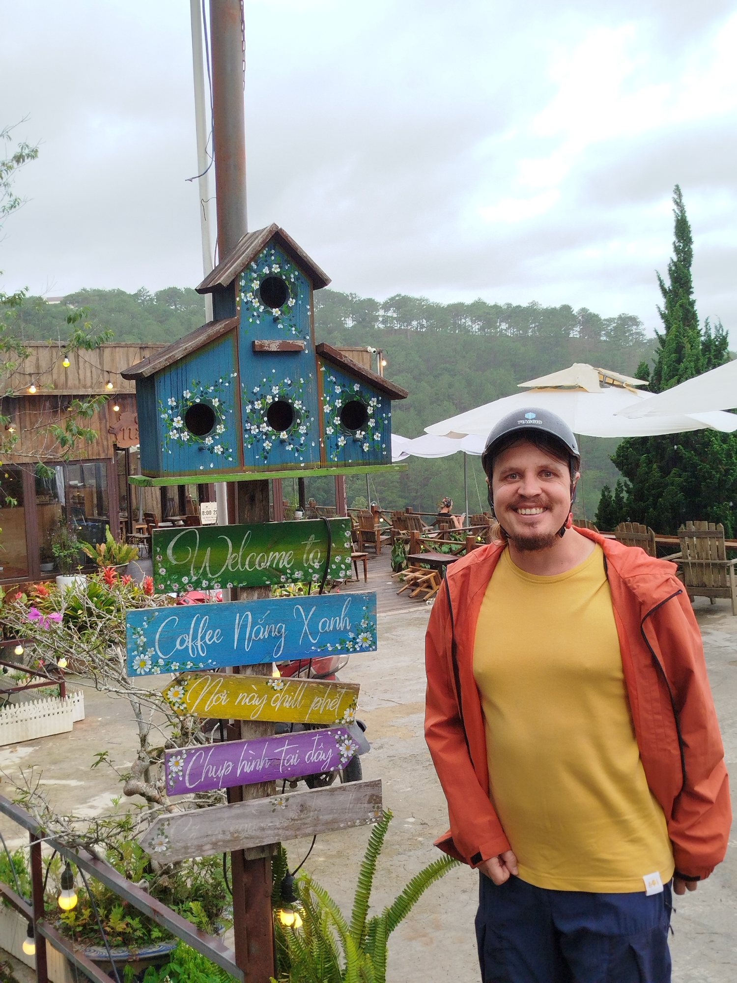 The image shows a man standing next to a signpost in what appears to be a tourist area in Da Lat, Vietnam. The man is wearing a yellow t-shirt, an orange jacket, dark blue pants and a black helmet, and smiling. The signpost is decorated with colorful flowers and features a blue birdhouse on top. The signs are painted in Vietnamese script, likely indicating directions to different attractions. One sign reads "Welcome to" in English, while another reads "Coffee Nang Xanh". Other signs say "Nơi này chill phet" and "Chụp hình tại đây." The area behind the signpost appears to be a cafe or restaurant with wooden furniture and umbrellas. In the background, there is a hilly landscape with trees and an overcast sky.