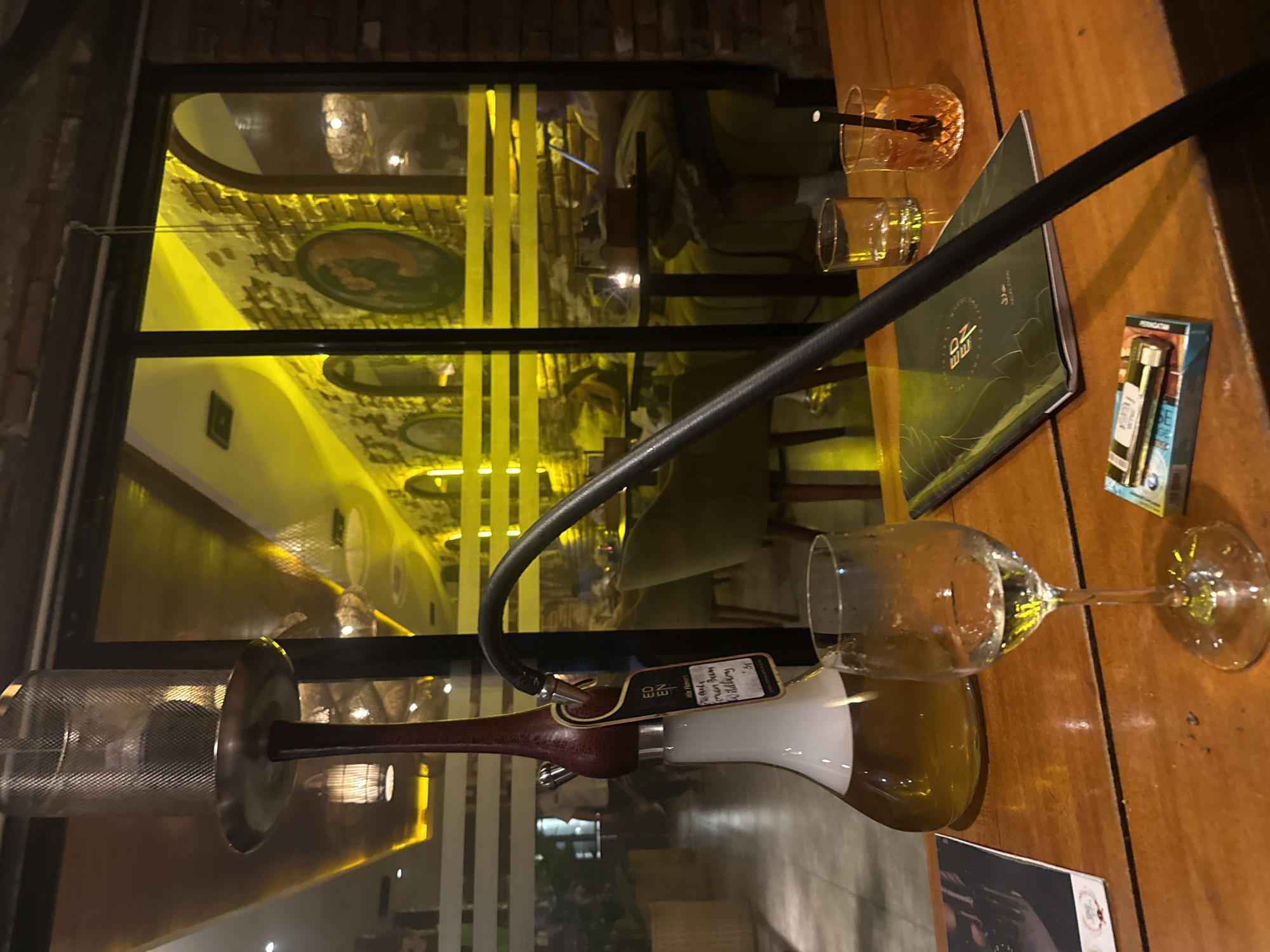 The image captures a nighttime scene, appearing to be indoors possibly in a restaurant or lounge, located in North Kuta, Indonesia. On a wooden table, there's a hookah, a half-filled wine glass, and several drinking glasses along with a menu and pack of cigarettes. The background is a reflection of a building with illuminated interiors, marked by bright yellow lights.