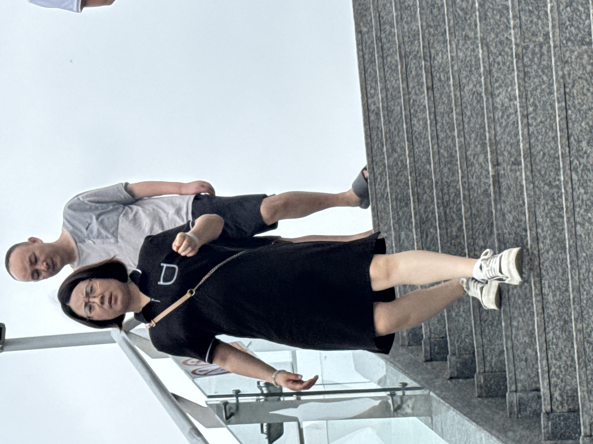 An outdoor, eye-level shot captures a woman and a man descending a grey stone staircase, possibly in the Shapingba District of China. The woman, in the foreground, wears a black short-sleeved dress and white athletic shoes. She has short dark hair, wears glasses, and carries a light-colored crossbody bag. A silver bracelet adorns her left wrist. Behind her, the man wears a grey t-shirt and dark shorts, and grey slide sandals. The background consists of a bright, overcast sky and the glass railing of the staircase. The stone steps are textured with grey and white flecks.