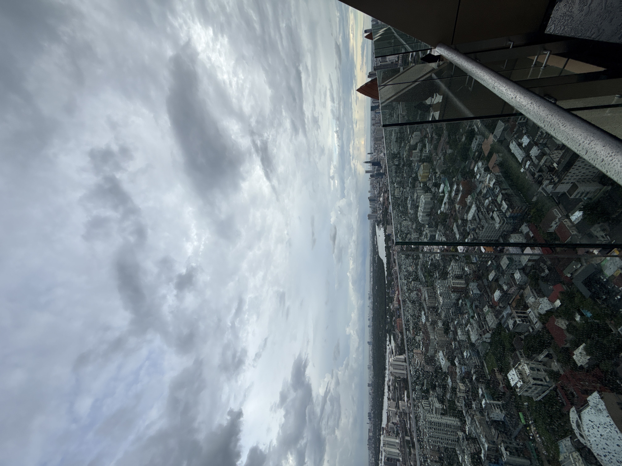 This is a high-angle, wide shot of a sprawling cityscape in Bangkok, Thailand, under a heavily overcast sky. The foreground features a balcony with a glass railing, exhibiting numerous water droplets, suggesting recent rain. The balcony is attached to a modern building with a dark, angular facade.
Beyond the balcony, the city unfolds with a dense collection of buildings, ranging from high-rise apartments and commercial towers to smaller residential structures. The architecture is varied, with a mix of modern glass-fronted buildings and older, more traditional-looking houses. Greenery is interspersed throughout the urban landscape, indicating parks or tree-lined streets.
In the mid-ground, a wide river, likely the Chao Phraya River, curves through the city, separating different districts. The far distance shows a hazy skyline dotted with more skyscrapers, characteristic of a major metropolitan area. The overall atmosphere is muted and somber due to the cloudy conditions, which also obscure any clear view of the horizon or distant mountains. No people are visible in the image. The scene captures the dense, vibrant, and dynamic nature of Bangkok, even under inclement weather.