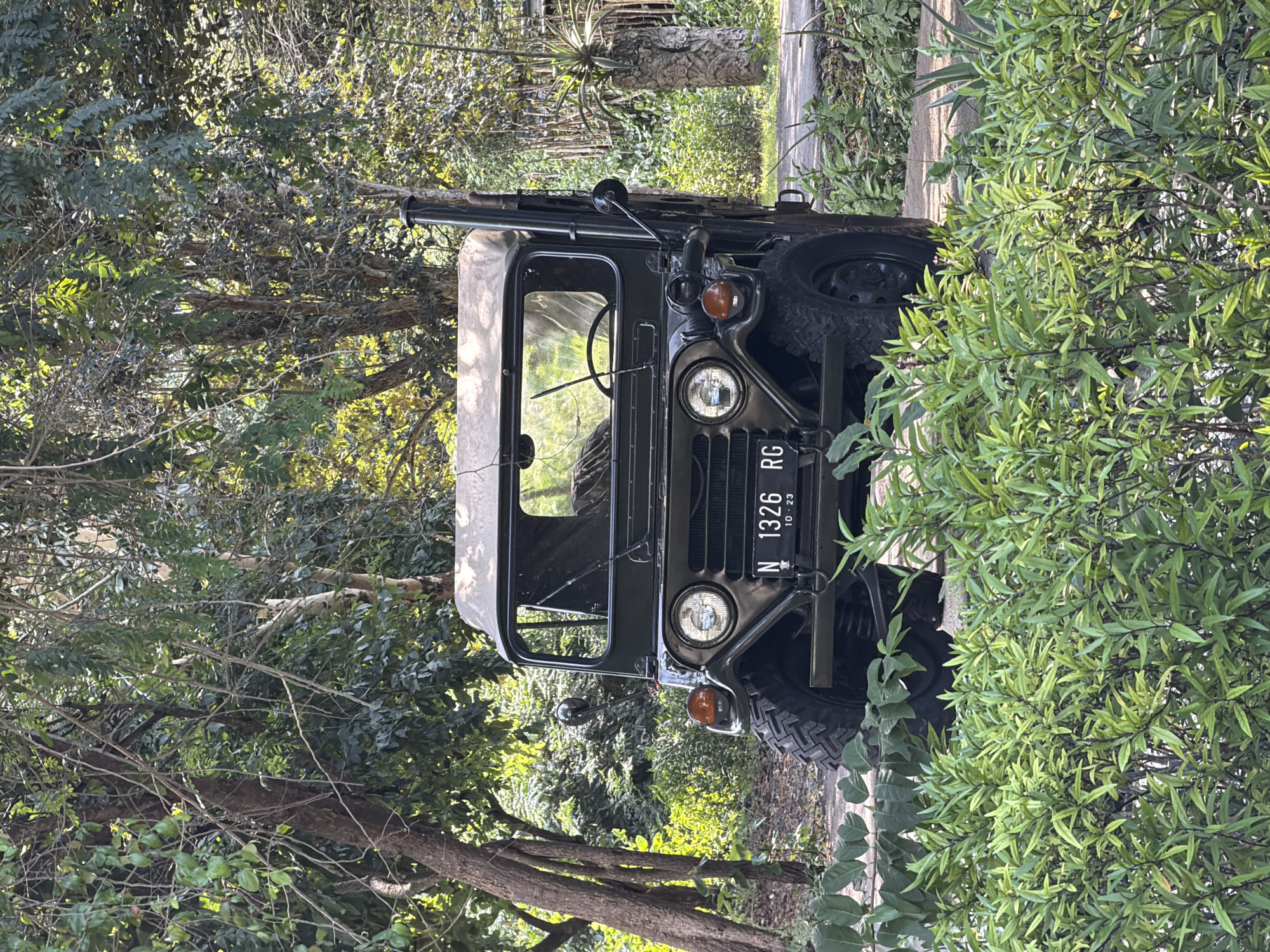 The image shows a dark green jeep, possibly an older model, parked amidst dense foliage, likely on a roadside or within a park in Indonesia. The license plate reads "N 1326 RG". The vehicle is surrounded by trees and bushes, obscuring much of the background. A paved surface is visible near the front tire of the vehicle.
