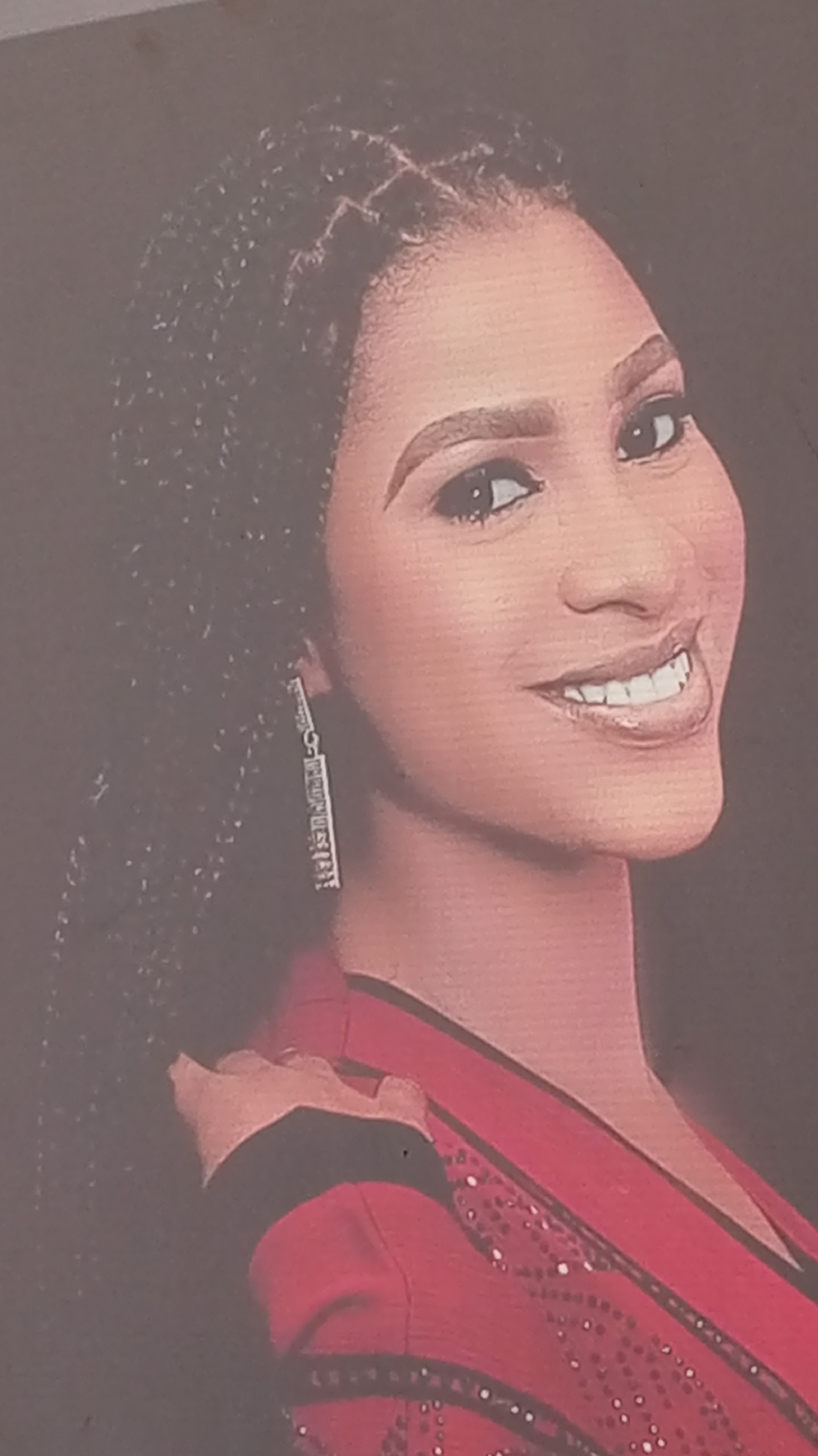 A portrait of a woman with braided hair, wearing a red top and a dangling earring is displayed. The location, Sabon Tasha, Nigeria, is not discernible from the image itself. The image is a close-up shot focusing on the woman's face and upper body.
