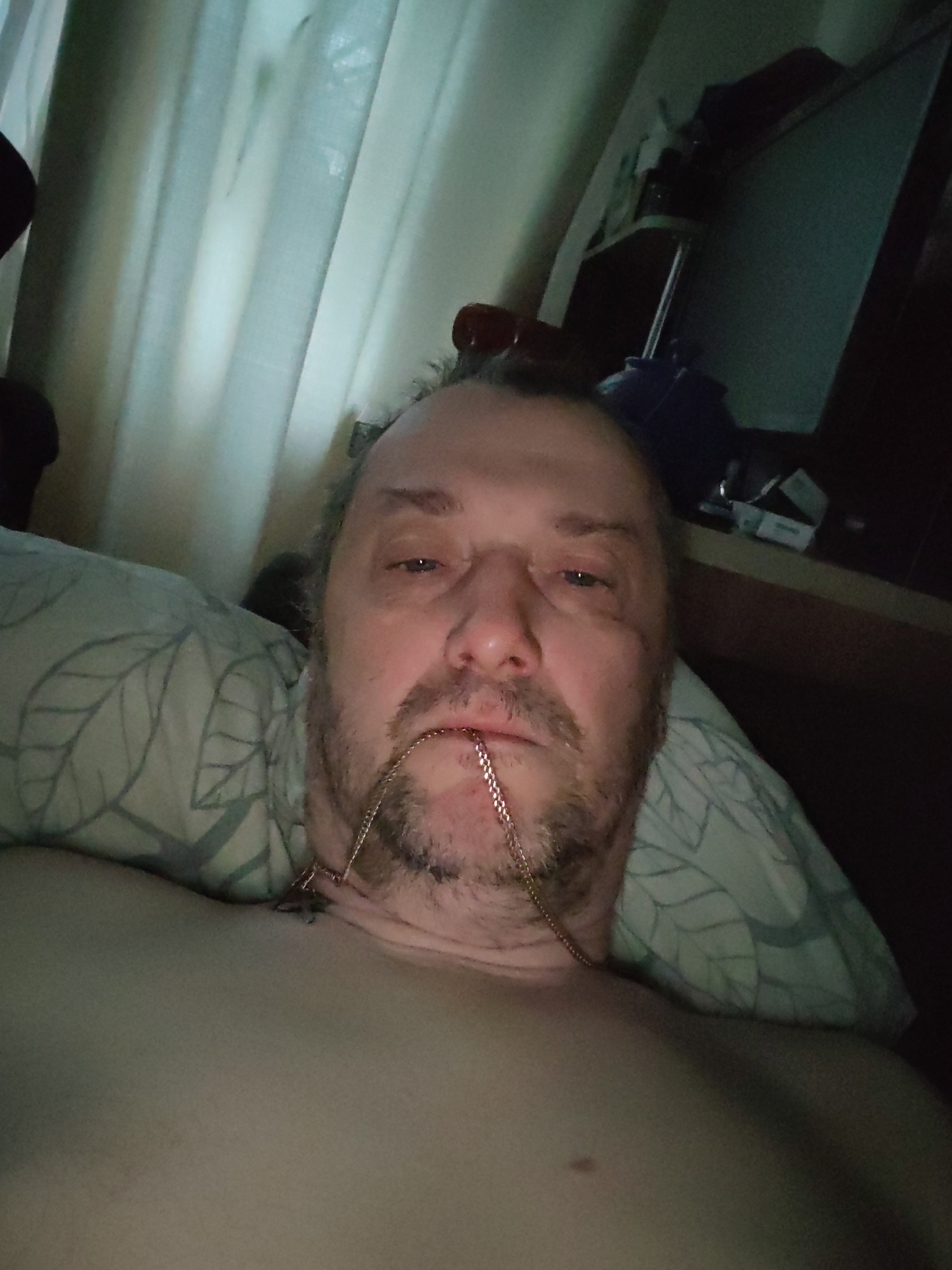 The image shows a middle-aged, unshaven man lying in bed, with a gold chain in his mouth. The background includes a pillow with a leaf pattern, a window covered by curtains, and a desk with objects on it, potentially electronic equipment and packaging. The man is topless. There is no evidence to indicate the location of Chaikovsky, Russia.