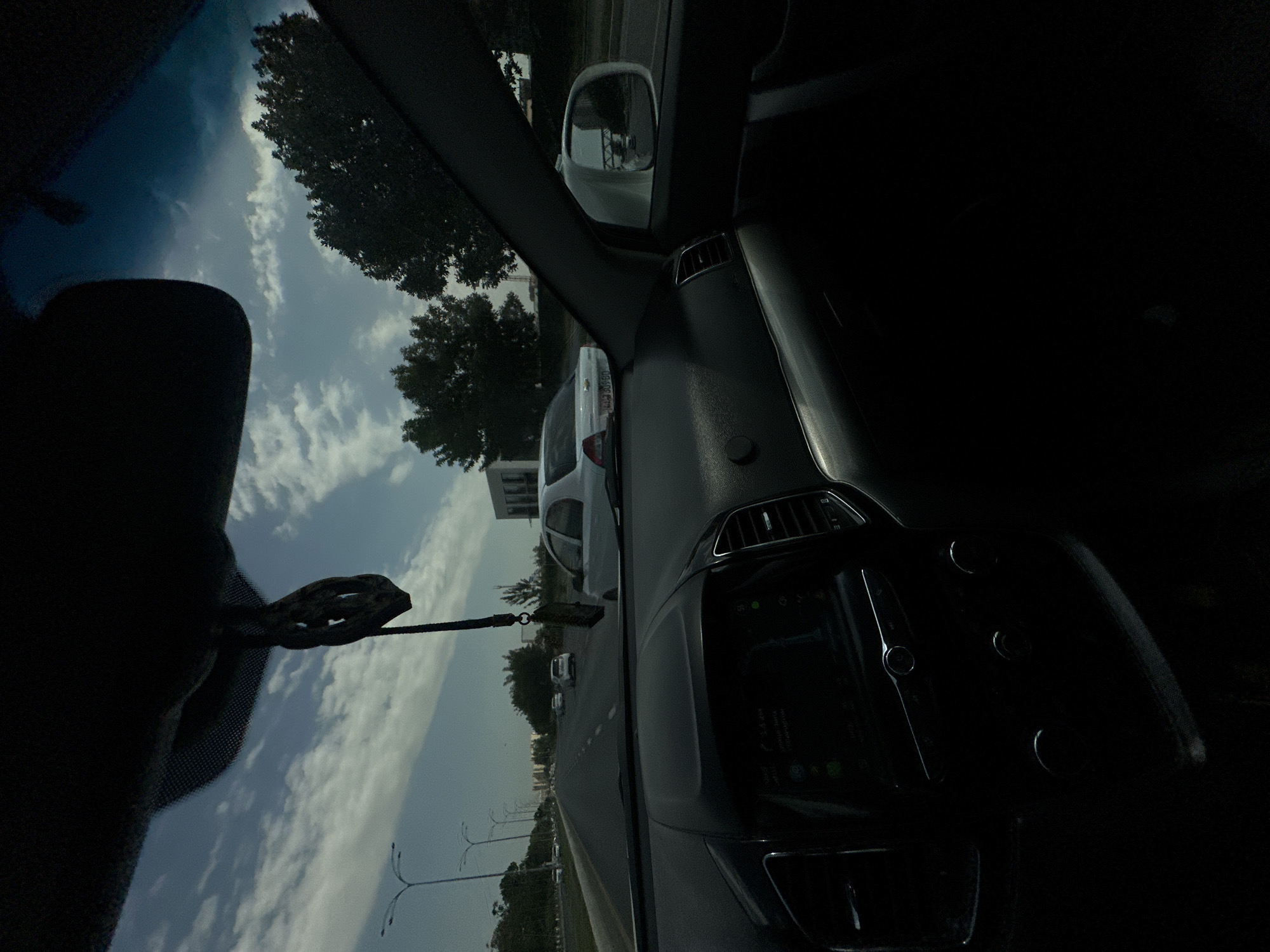 The image is taken from inside a car, looking forward through the windshield. The car is on a multi-lane road, with other vehicles visible ahead. The sky is partly cloudy. Trees and buildings are visible on either side of the road. The dashboard of the car is in the foreground, with a navigation screen and controls visible. A hanging car air freshener is visible in the center of the windshield. The scene appears to be a typical urban or suburban environment.
Based on the license plate on the white car in front, which reads "09 305 GD", and its typical format, this location is likely Uzbekistan. Based on the presence of streetlights and the overall urban infrastructure, the image may have been taken in Tashkent, the capital city of Uzbekistan.