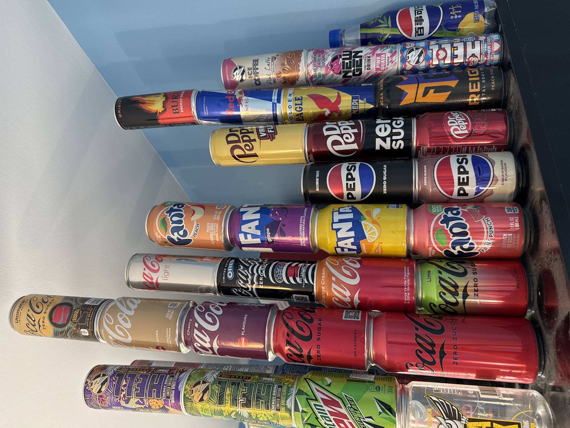 The image shows a collection of soda and energy drink cans and bottles stacked in a pyramid-like arrangement, presumably on a shelf. The cans are various brands and flavors, including Coca-Cola (classic, zero sugar, cherry, orange cream, lime, vanilla, light, ultimate limited edition), Pepsi (classic, zero sugar, cream soda), Fanta (peach, grape, lemon, fruit punch), Mountain Dew, and various energy drink brands such as Burn, Red Bull, Golden Eagle, Reign, New Gen, and a Japanese Pepsi variant. There are no people visible in the image. The location is not discernible based on the information in the image, therefore Burgas, Bulgaria cannot be confirmed as the location.