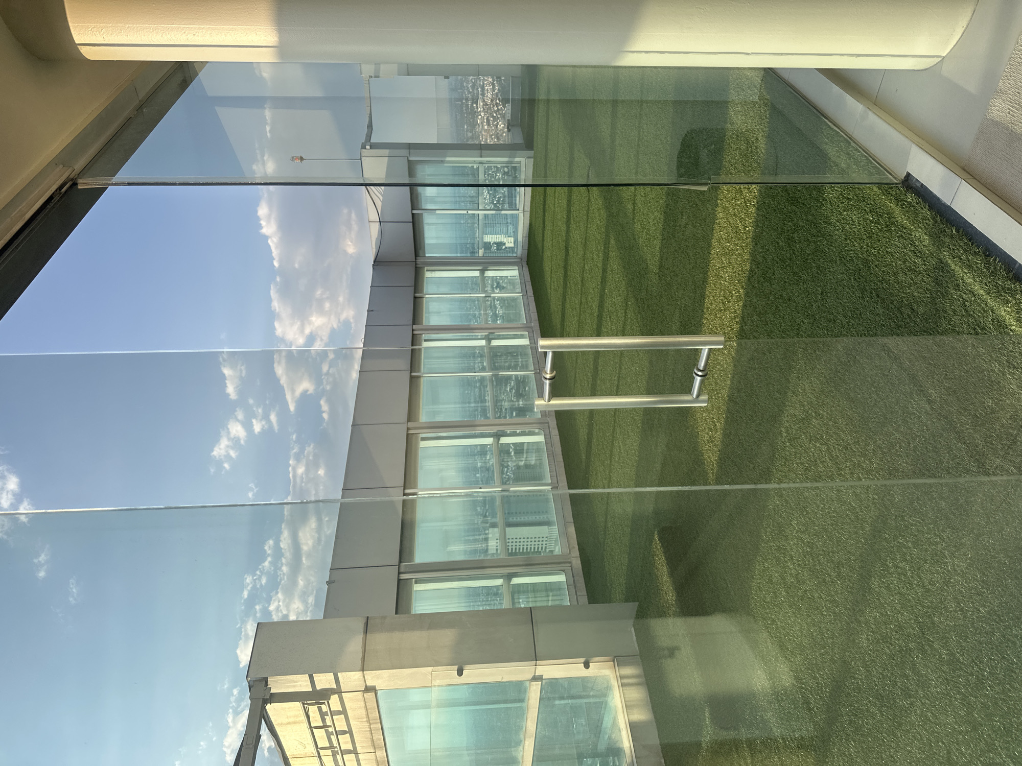 The image shows a view from inside a building in what appears to be San Bernardino Tlaxcalancingo, Mexico. There is a glass wall providing the view. The exterior features astroturf. Reflections in the glass show a blue sky with white clouds and adjacent buildings. There's a metal handle attached to the glass. A city landscape is visible in the distance. The interior of the building is partially visible above the glass.