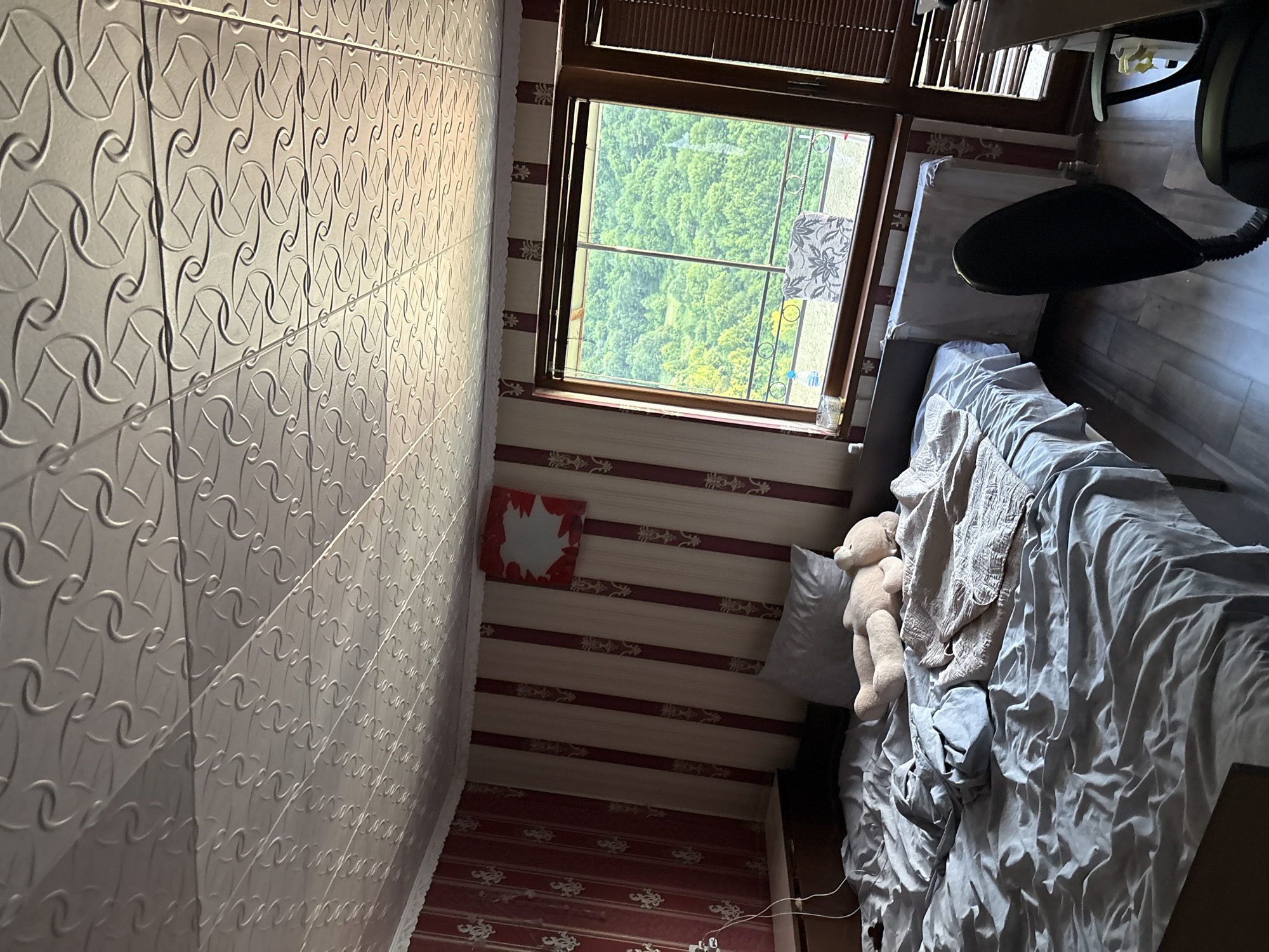 The image depicts a bedroom with a bed, a window, and a ceiling. The bed is unmade, covered with gray sheets and a tan blanket, with a teddy bear lying on it. A gray pillow is visible at the head of the bed. The walls are wallpapered with alternating vertical stripes of red and cream, featuring a decorative gold pattern. Above the walls, the ceiling is a white textured surface with a repeating geometric pattern. A framed artwork, predominantly red with a white center, hangs on the wall. The window offers a view of a lush green forest or hillside. To the right of the window, a radiator is visible, and blinds cover part of the window. In the foreground, a portion of a black office chair is visible. The scene appears to be an interior residential space, possibly located in Redina, Bulgaria, based on the prompt's context.