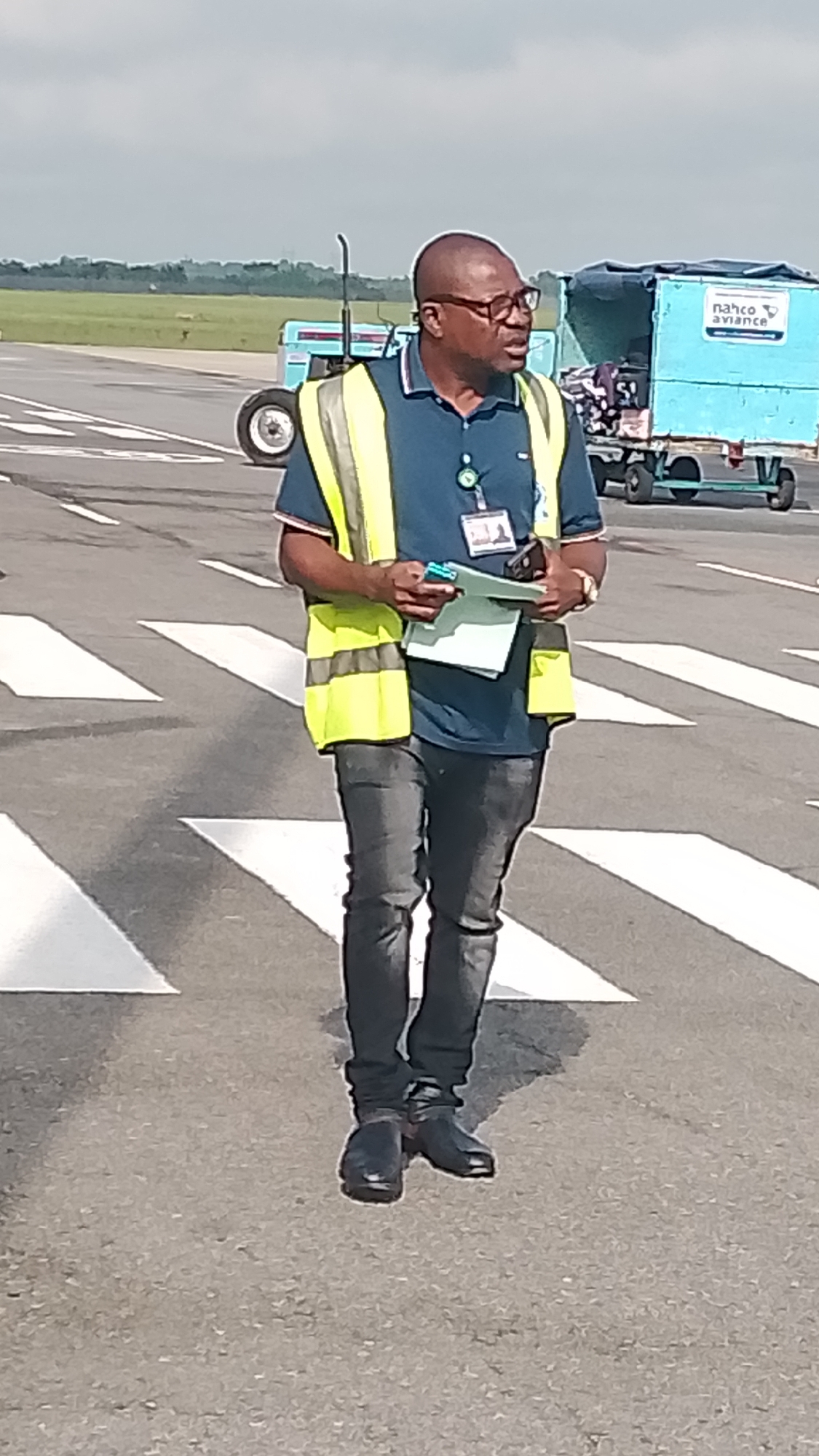 A medium shot captures a bald Black man wearing glasses, a dark blue polo shirt, a bright yellow high-visibility vest, and dark jeans on an airport tarmac. He stands on a crosswalk with white stripes, looking to his right and holding papers and a pen. Behind him, a light blue service cart with the logo "NAHCO AVIANCE" is visible, along with a green field and a hazy sky. The scene appears to be in Orerokpe, Nigeria.