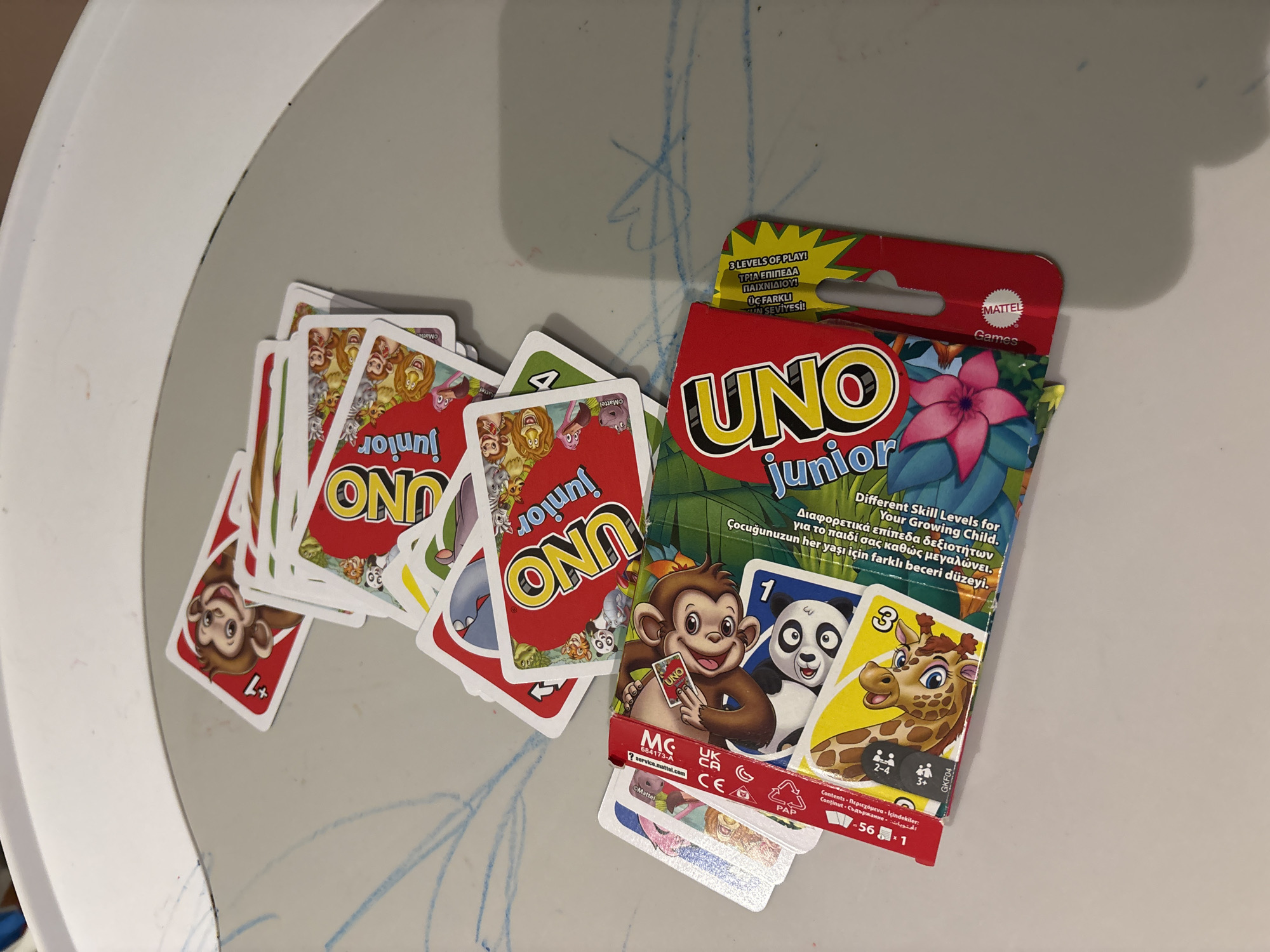 The image shows a collection of UNO Junior playing cards scattered on a light gray surface, which appears to be a table. The cards are brightly colored, featuring various cartoon animal illustrations and the UNO logo. Some cards are fanned out, while others are stacked or partially obscured. One card prominently displays a monkey with "+1" text. Another card, part of a red box, shows a panda and a giraffe. The box also has text in English and possibly other languages, indicating "3 LEVELS OF PLAY!" and "Your Growing Child." The cards appear to be new or in good condition. The background is a white, curved surface, possibly the edge of a table or tray. There are faint blue scribbles on the gray surface, suggestive of children's drawings. The scene does not provide any information that would allow for the identification of a specific location within Dubai, United Arab Emirates, or any people or actions taking place.