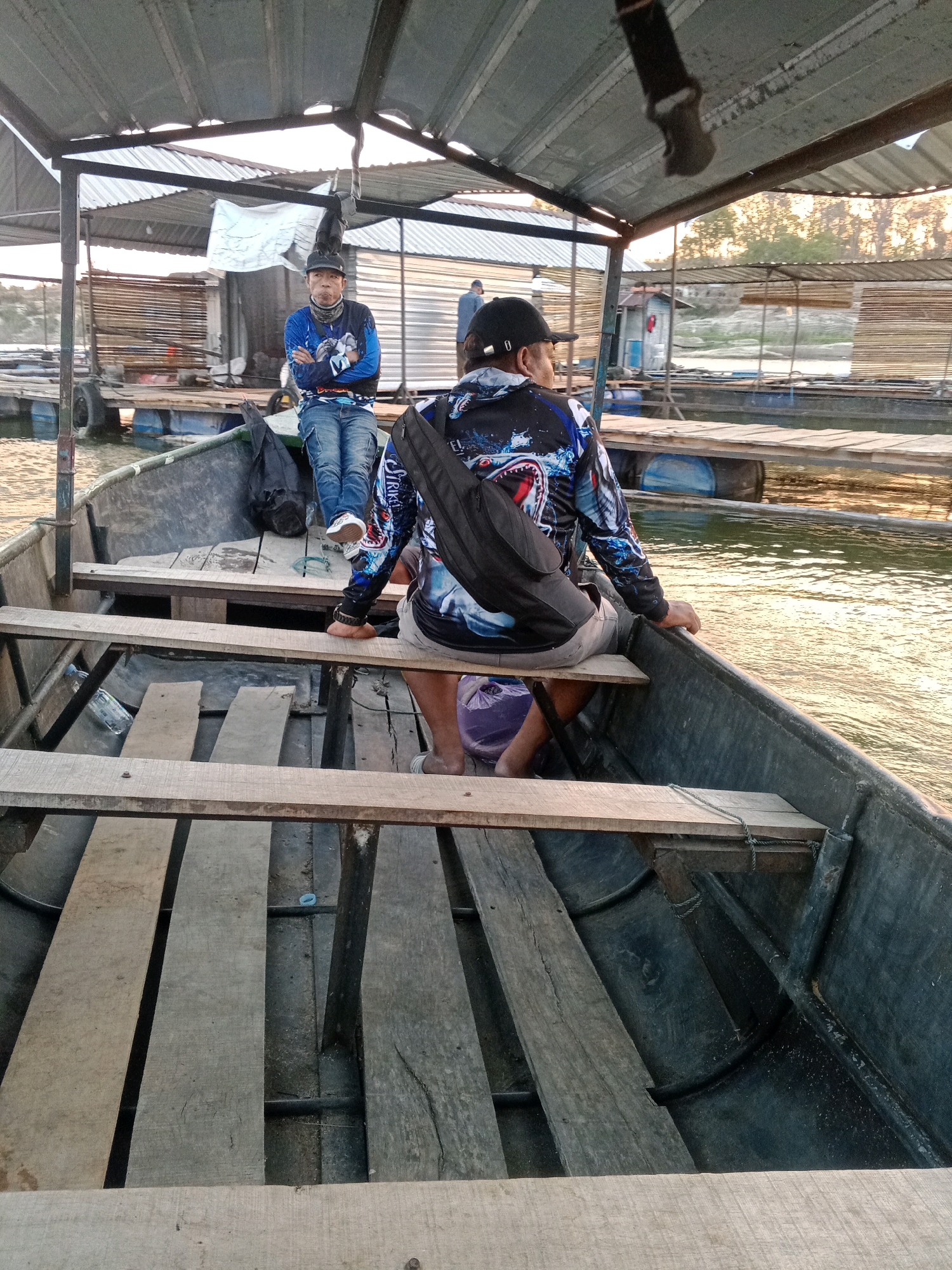 The image depicts two individuals in a boat docked at what appears to be a fish farm in Sendang, Indonesia. The boat is made of dark grey plastic with wooden benches. One person, wearing a blue long-sleeved shirt and jeans, sits facing the camera with their arms crossed. The second person, wearing a black long-sleeved shirt with a shark graphic and shorts, is seated with their back to the camera, a black bag slung over their shoulder. In the background, a structure with a corrugated metal roof is visible, along with wooden platforms and cages, typical of a fish farming operation. The water surrounding the boat is calm and reflects the warm light of what appears to be late afternoon or early evening.