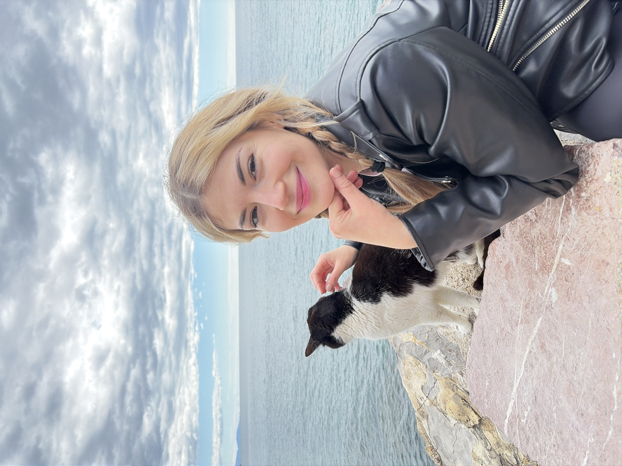 A woman with blonde hair braided on one side stands on a stone ledge overlooking the Adriatic Sea, petting a black-and-white cat. The location appears to be a coastal area near Budva, Montenegro. The sky is overcast. The woman is wearing a black leather jacket and dark leggings. She is smiling. The cat is standing on the ledge with the ocean visible in the background.