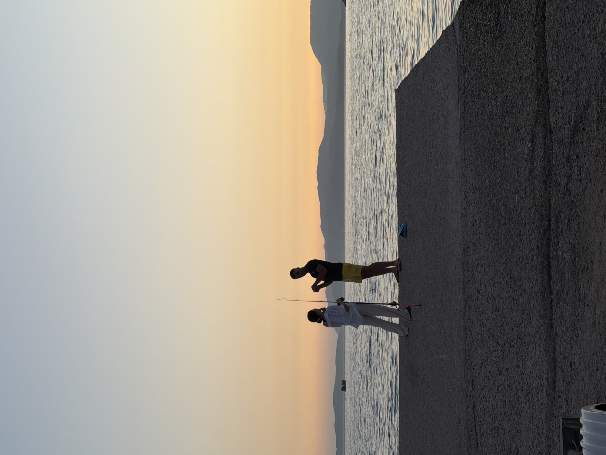 This is a full-frame, eye-level, outdoor shot of a pier in the Municipal Unit of Alyzia, Greece, at dusk. The sky is a gradient of pale blue to warm orange, with a silhouette of mountains in the distance. The sea is calm with gentle ripples. Two figures are silhouetted on the pier, a woman and a man, both facing the sea. The woman, on the left, is wearing a white shirt and light-colored pants, holding a fishing rod. The man, on the right, is wearing yellow shorts and a dark shirt, holding what appears to be fishing line in his hands. They are positioned approximately two feet apart. A small boat is visible in the distance on the left. The pier is concrete with a textured surface. A blue object, possibly a bag, lies on the pier to the right of the figures.