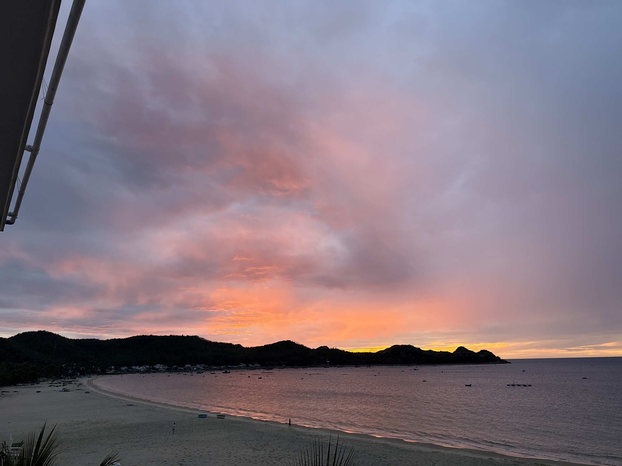 The image depicts a serene sunset over a coastal area in Song Cau, Vietnam. The sky is a canvas of vibrant colors, transitioning from soft pinks and oranges near the horizon to muted purples and blues higher up. Wispy clouds catch the last rays of sunlight, creating a dramatic and picturesque scene.
In the foreground, a sandy beach stretches along the coastline, appearing mostly empty except for a few small, dark figures that are likely people walking. Some small boats are visible on the beach, suggesting recent activity or preparation for the evening.
The middle ground is dominated by the calm sea, reflecting the colors of the sky. Several small boats are scattered across the water, some appearing to be anchored. A cluster of small structures, possibly part of a village or settlement, lines the edge of the bay.
In the background, a dark, hilly silhouette forms the horizon. These hills are covered in dense vegetation, creating a natural backdrop to the coastal landscape. The overall impression is one of tranquility and natural beauty, characteristic of a peaceful evening in a coastal Vietnamese town.