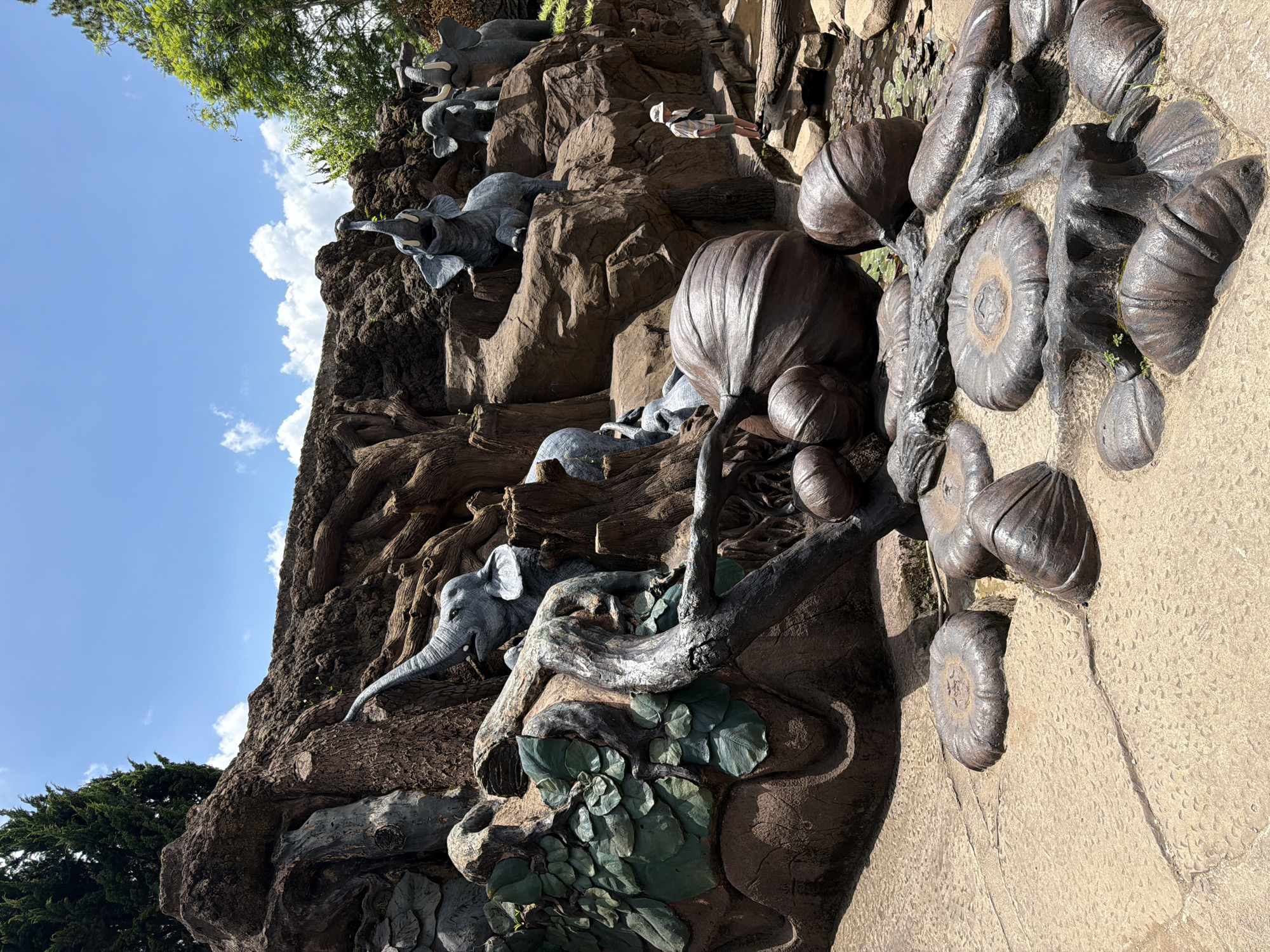 The image depicts a scenic outdoor installation, likely a tourist attraction, featuring several sculpted elephants integrated into a rocky, tree-root-like landscape. The elephants are grey and vary in size and pose; some are depicted with their trunks raised. In the foreground, large, dark brown, stylized seed-like or fruit-like sculptures are arranged on a paved surface. A small stream with lily pads is visible in the background, and a person wearing a hat and shorts stands on a rock near the water. The overall setting is naturalistic with lush green trees and a blue sky with scattered clouds overhead. This scene is located at the Da Lat Flower Garden in Da Lat, Vietnam.