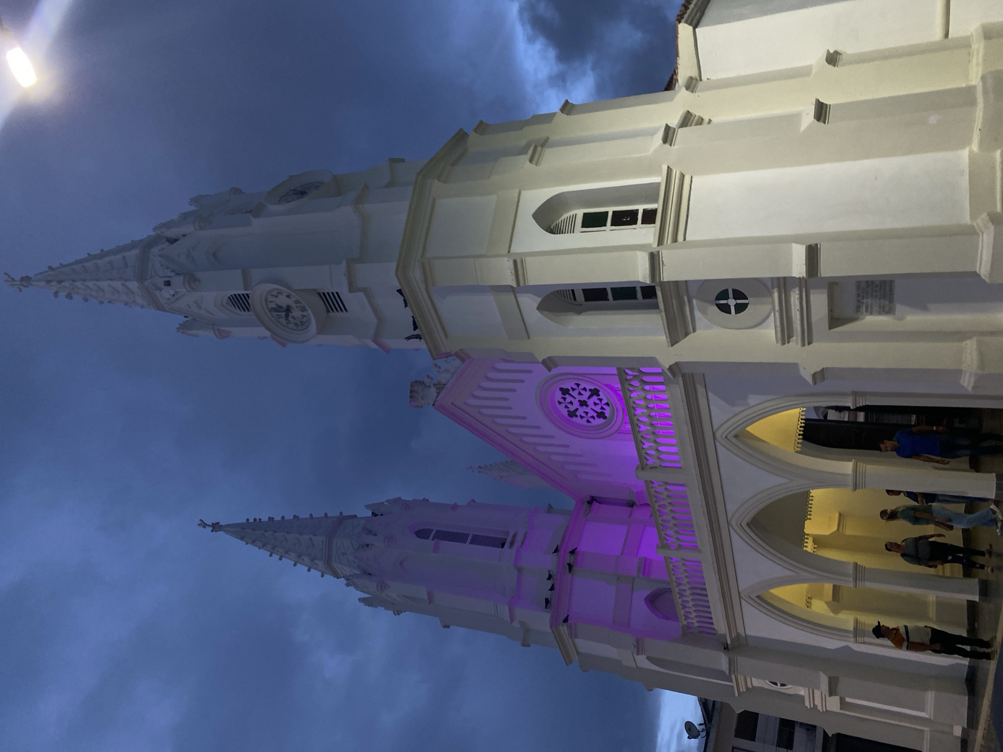 The image shows the Iglesia San Juan Evangelista, a white church in Juan Griego, Venezuela. The church has a tall spire with a clock on one side. The building is illuminated with white and purple lights. Several people are visible under the arched entryways of the building. The sky is a dark blue.