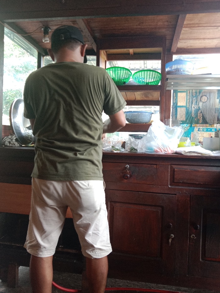 A man in a dark green t-shirt, beige shorts, and a dark baseball cap stands facing away from the viewer in a wooden cart. Above the counter are shelves with plastic baskets. The man appears to be working at the counter, with plastic bags and various items visible. The cart has a window, allowing natural light to illuminate the scene. Below the counter are drawers with keys visible in the keyholes.