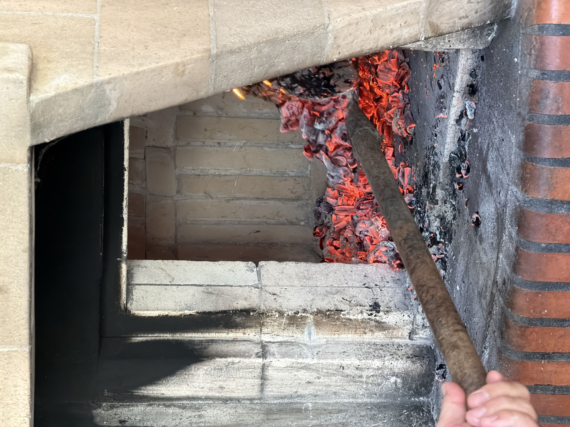 The image shows the interior of a brick oven, likely a wood-fired oven. Inside, there are burning embers, glowing red and gray, being moved or manipulated with a long wooden pole held by a person whose hand is visible in the lower right corner of the frame. The oven's interior is made of brick, showing signs of soot and heat discoloration. The opening of the oven is framed by light-colored bricks or stone, creating a contrast with the darker interior. The location is Vila Real, Portugal.
