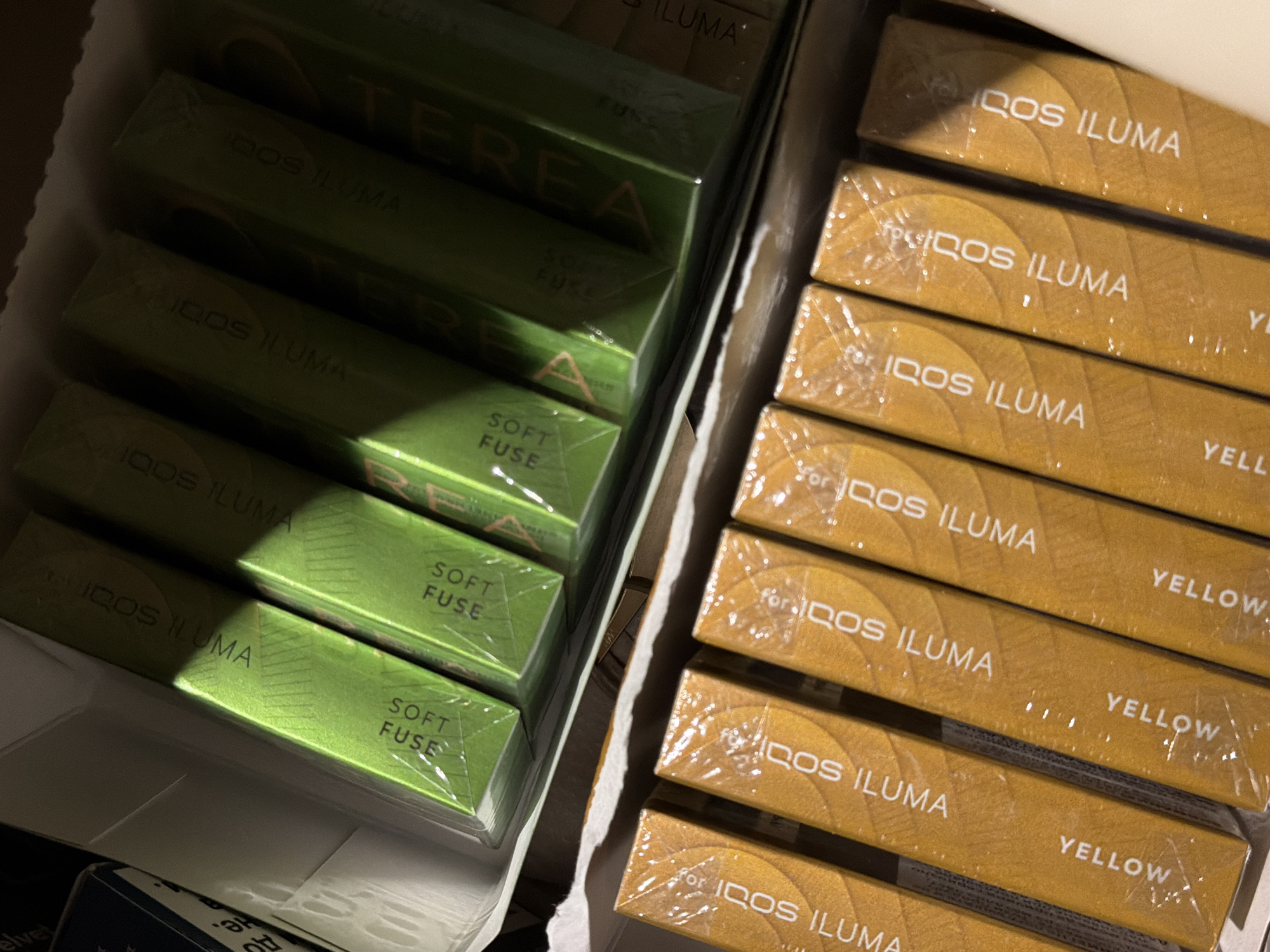 The image shows two rows of packaged tobacco sticks, likely for use with an electronic device. The top row contains green packages with "IQOS ILUMA SOFT FUSE" printed on them, along with the brand name "TEREA". The bottom row contains yellow packages with "IQOS ILUMA YELLOW" printed on them. The packages are arranged in a box and appear to be factory-sealed. The lighting suggests the image was taken indoors, possibly in a retail or storage setting. There are no people or specific environmental details that would indicate a location in Burgas, Bulgaria.