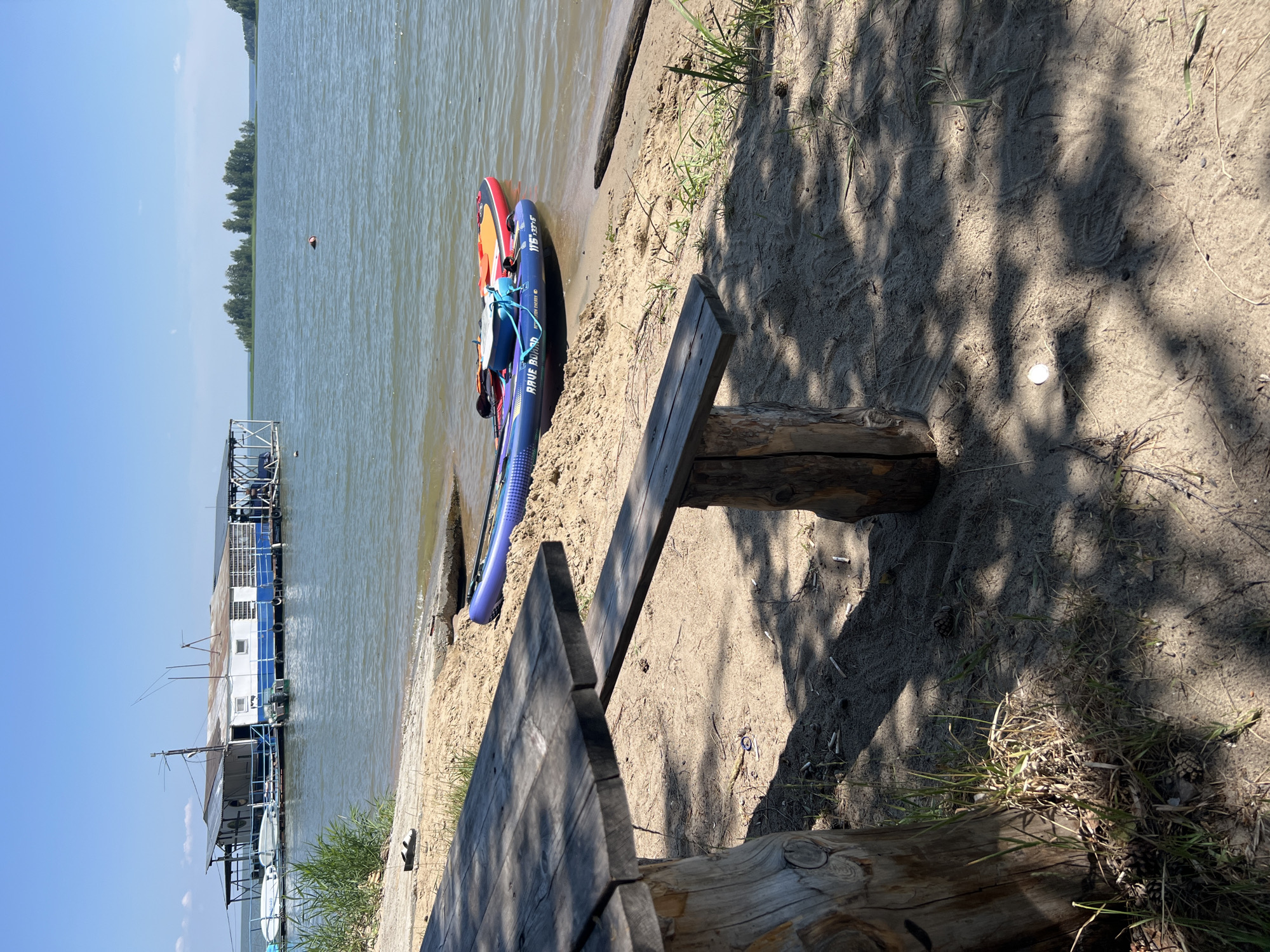 A wooden picnic table and bench sit on a sandy shore next to a calm body of water under a clear blue sky. Two stand-up paddleboards, one blue and one orange, are partially submerged in the water near the shore. In the background, a long, white and blue floating structure with a metal roof is moored in the water. Small islands with trees are visible in the distance. The scene appears to be a leisure area at Lake Borovoe, Russia, a popular tourist destination.