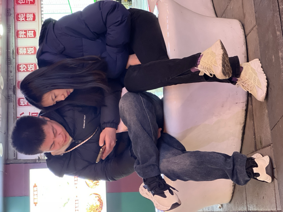Two young adults are seated side-by-side on a white, curved bench in an urban area of Yuzhong District, China. The male, positioned on the left, wears a black puffer jacket with "Come on! Start!" visible on the chest, a light-colored undershirt, and faded blue jeans. He holds a gold-colored smartphone in both hands, directing his gaze downwards at its screen with a focused expression. His right leg is crossed over his left, and he wears white and black sneakers. The female, seated to his right, wears a dark blue puffer jacket and black pants featuring small slits at the ankles. Her long dark hair falls forward as she leans slightly towards the male, also intently observing the smartphone screen. She wears cream-colored chunky sneakers over visible purple socks. The background reveals illuminated vertical menu boards displaying Chinese characters, which include "炒饭" (Fried Rice), "套饭" (Set Meal), "拌面" (Mixed Noodles), "炖罐" (Stew Pot), and "包子" (Buns), suggesting a food vendor or restaurant. The ground surface beneath the bench is a dark-colored pavement.