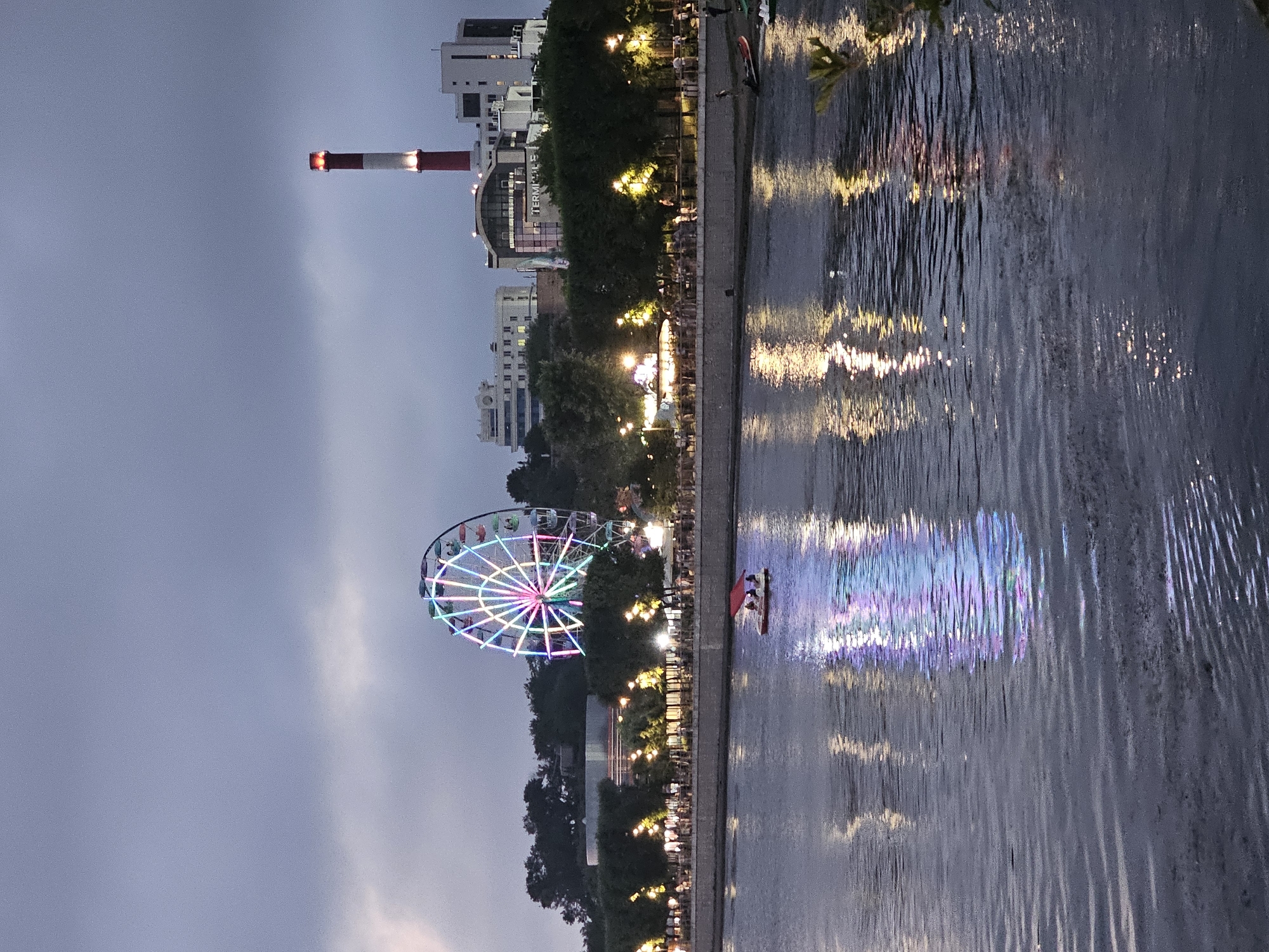 The image depicts a waterfront scene in Vladivostok, Russia, at dusk or early evening. A brightly lit Ferris wheel, adorned with a rainbow of colors, dominates the upper left portion of the frame, situated behind a line of trees. To the right of the Ferris wheel, a tall industrial smokestack with red and white bands stands out against the hazy sky, emitting a glow from its top. Several modern buildings are visible behind the trees and the Ferris wheel.
In the foreground, a wide body of water, likely a bay or river, reflects the lights from the shore and the Ferris wheel, creating shimmering patterns on its surface. A small red pedal boat with a canopy carrying at least two people is moving across the water. Along the waterfront, a promenade or park area is lined with trees and illuminated by numerous lampposts, with small groups of people visible walking or gathered. The overall atmosphere is one of a recreational area coming alive as evening falls.