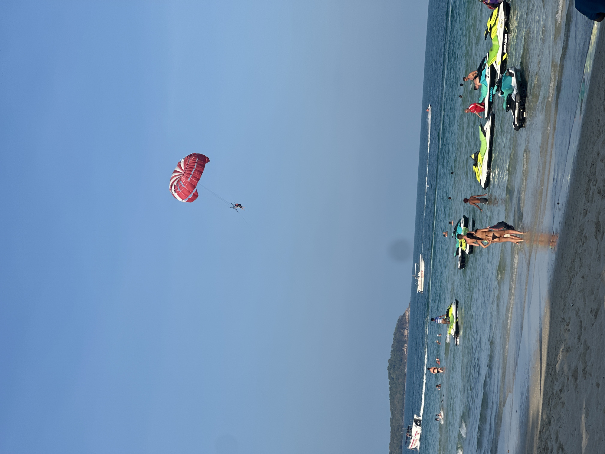 The image captures a beach scene in Baan Chaweng, Thailand, featuring a light blue sky, a shoreline with people, jet skis, and a parasailer in the sky.
The beach is populated with people, some standing in the shallow water, others sunbathing or interacting near jet skis parked along the shoreline. There are numerous jet skis, identifiable by their colorful design and brand names.
In the sky, a person is parasailing beneath a red and white parachute. The individual is suspended in the air, attached to the parachute, adding a dynamic element to the scene.
A boat is off shore with two levels of people, possibly a tour boat.