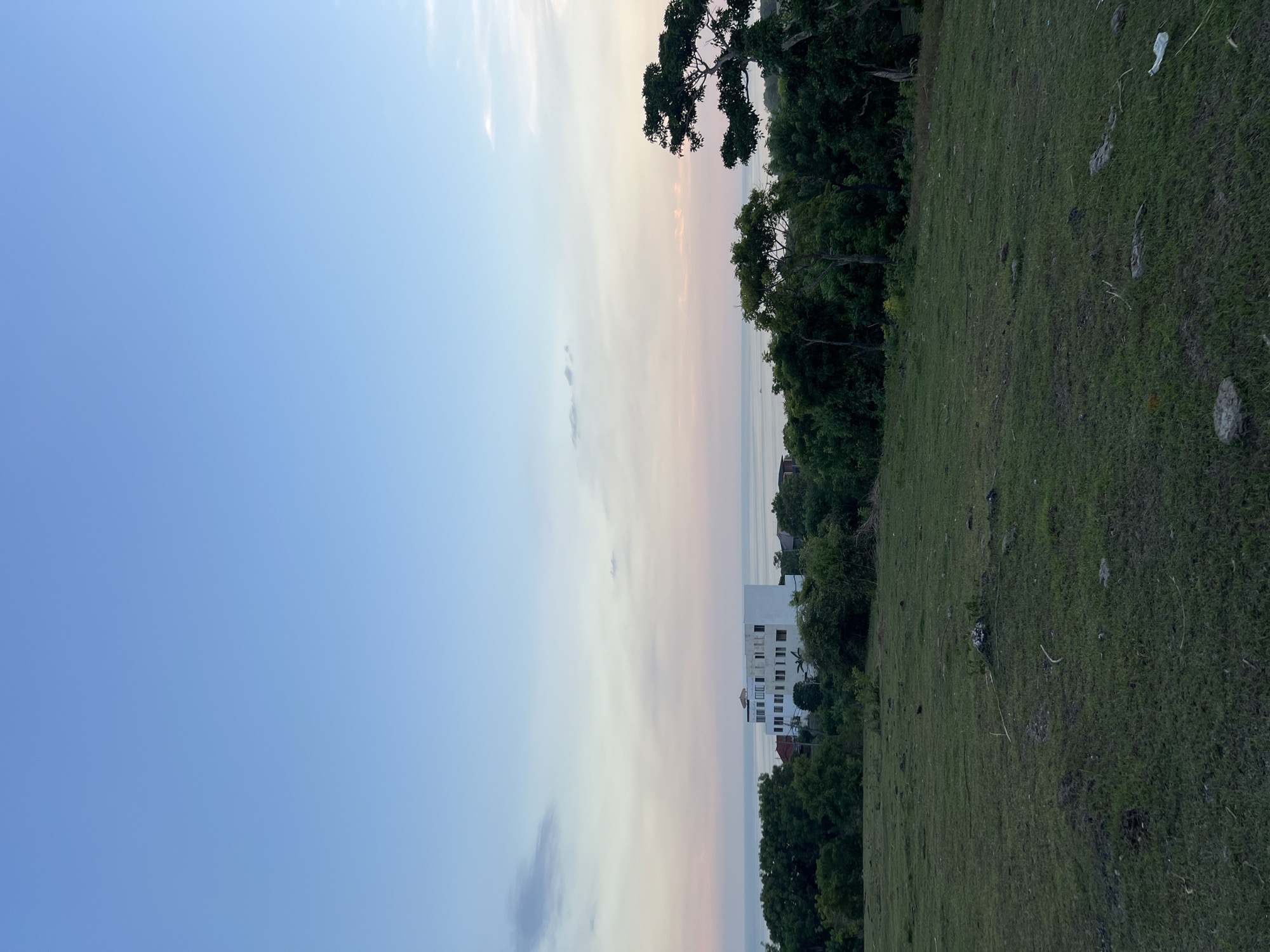 The image shows a landscape with a blue sky on the left, transitioning to a pinkish hue near the center, suggesting sunset or sunrise. On the right side, there is a green grassy area with trees. A white building is visible in the distance, partially obscured by the trees. The location is Unknown, Unknown.