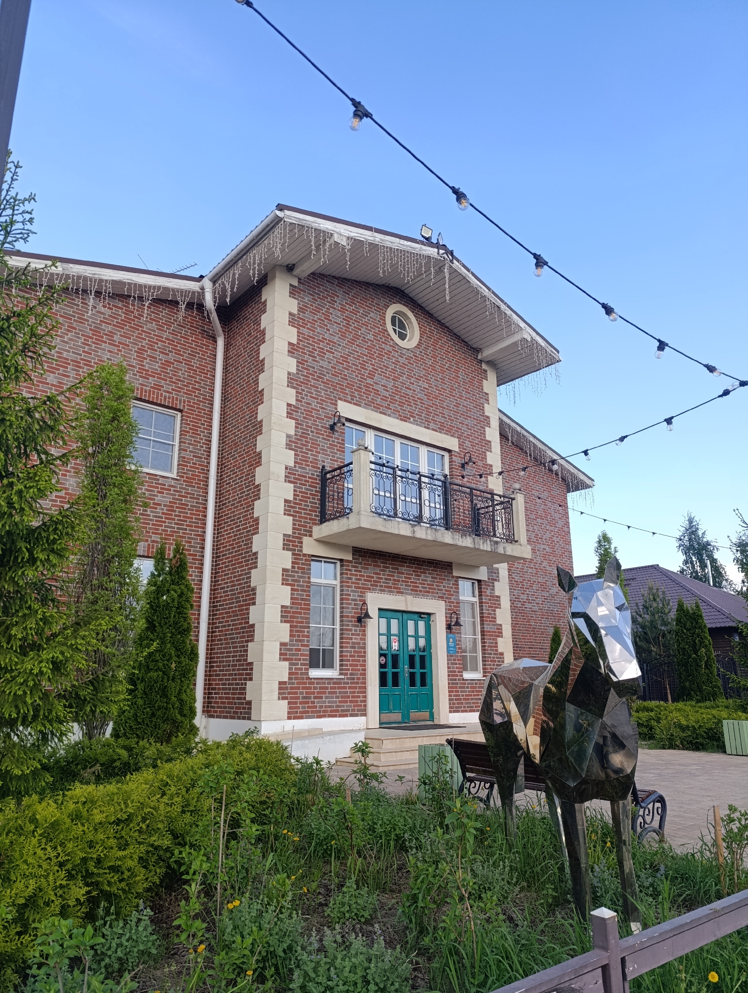 The image shows a two-story red brick building with white trim in Ivanovskoe, Russia. A green double door serves as the entrance. Above the door, a balcony with black metal railings is visible. A metallic, geometric horse statue stands in front of the building, surrounded by greenery. Overhead, string lights are strung diagonally across the scene. A bench is partially visible behind the statue. The sky is clear and blue. Other greenery, including trees and bushes, surrounds the building. Part of another building is visible to the right.